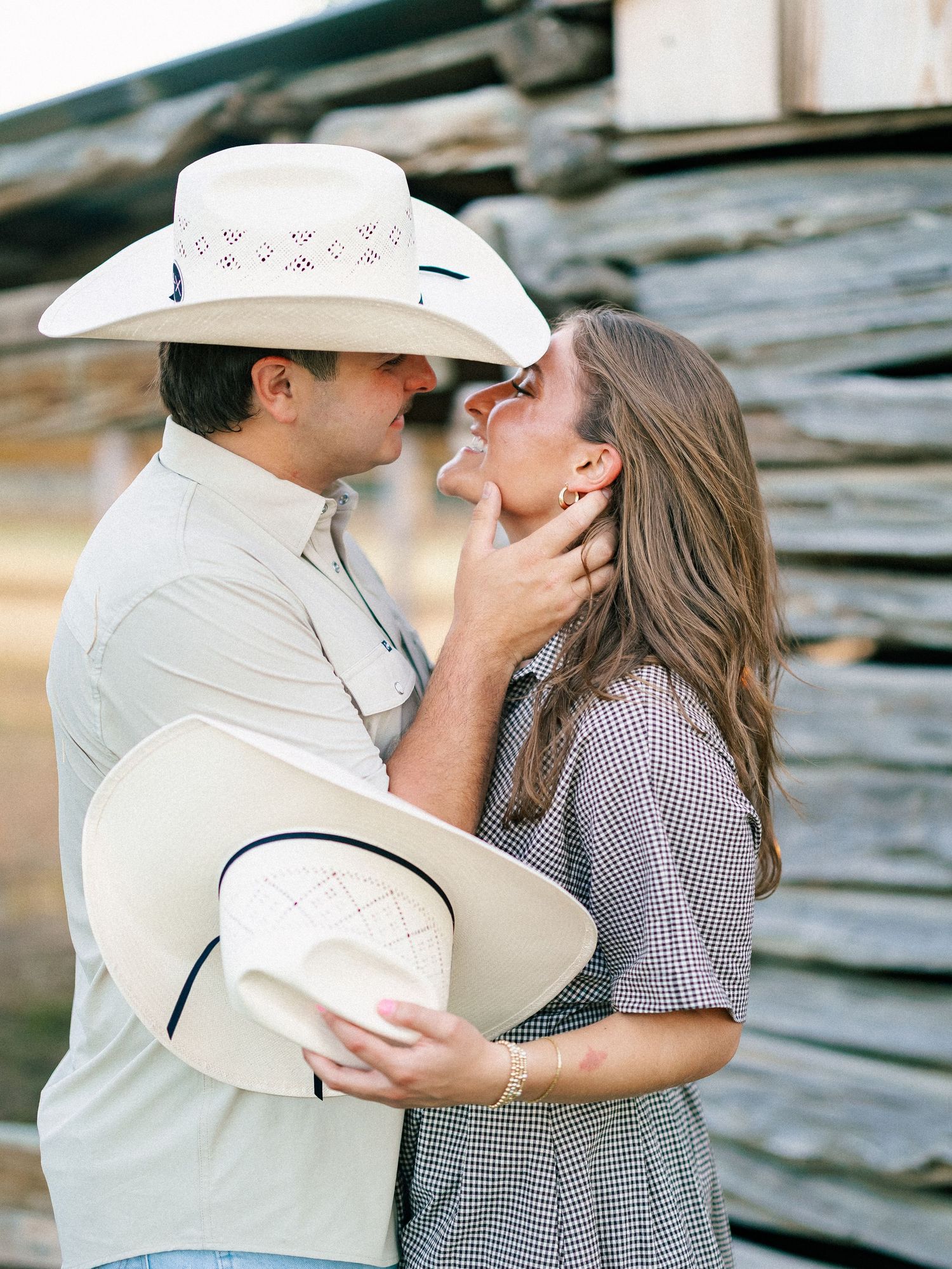 Couple in cowboy hats sharing a romantic moment near a rustic log wall during an outdoor engagement session.