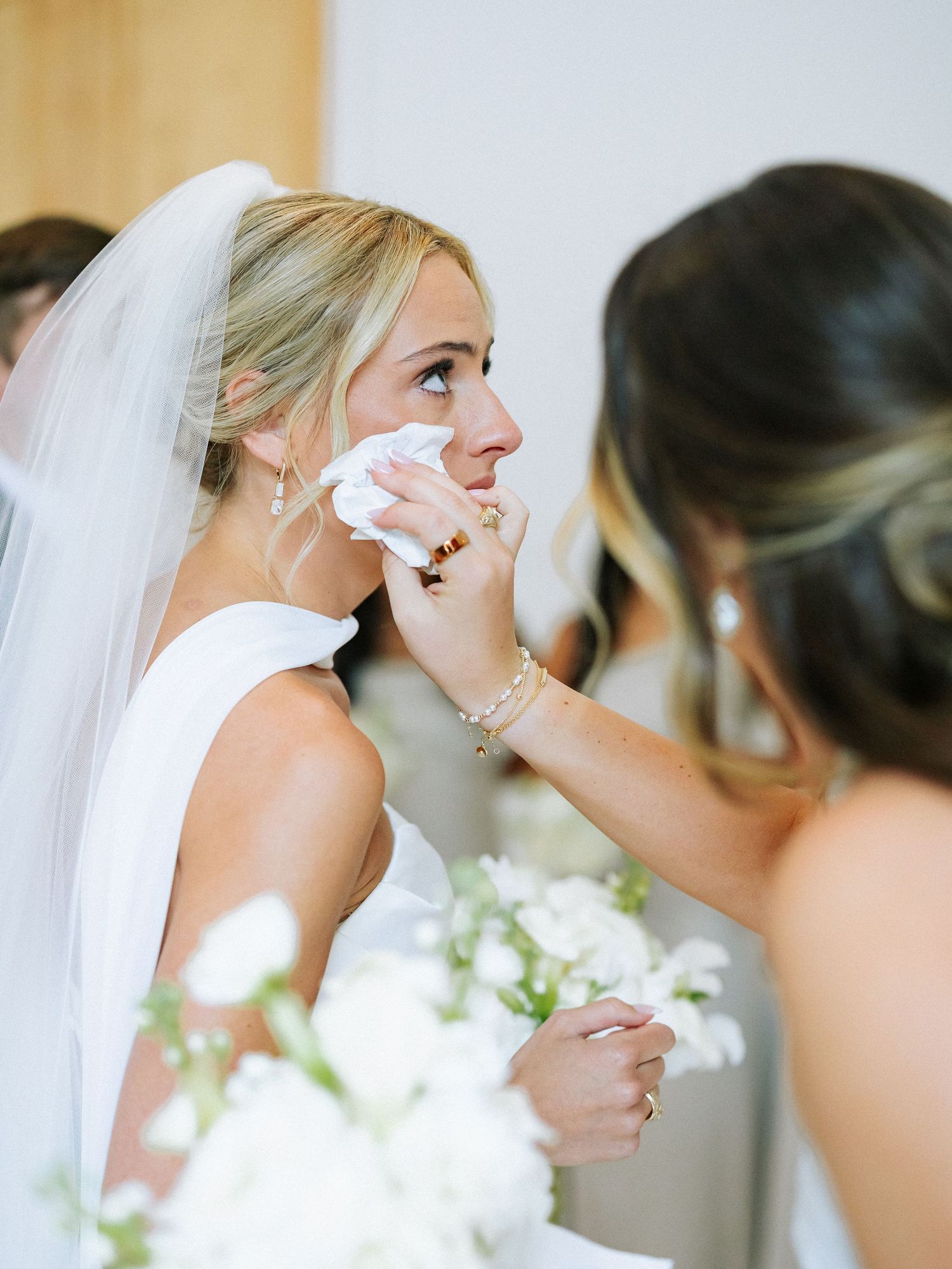 Bridesmaid gently dabs tears from emotional bride's face on wedding day.