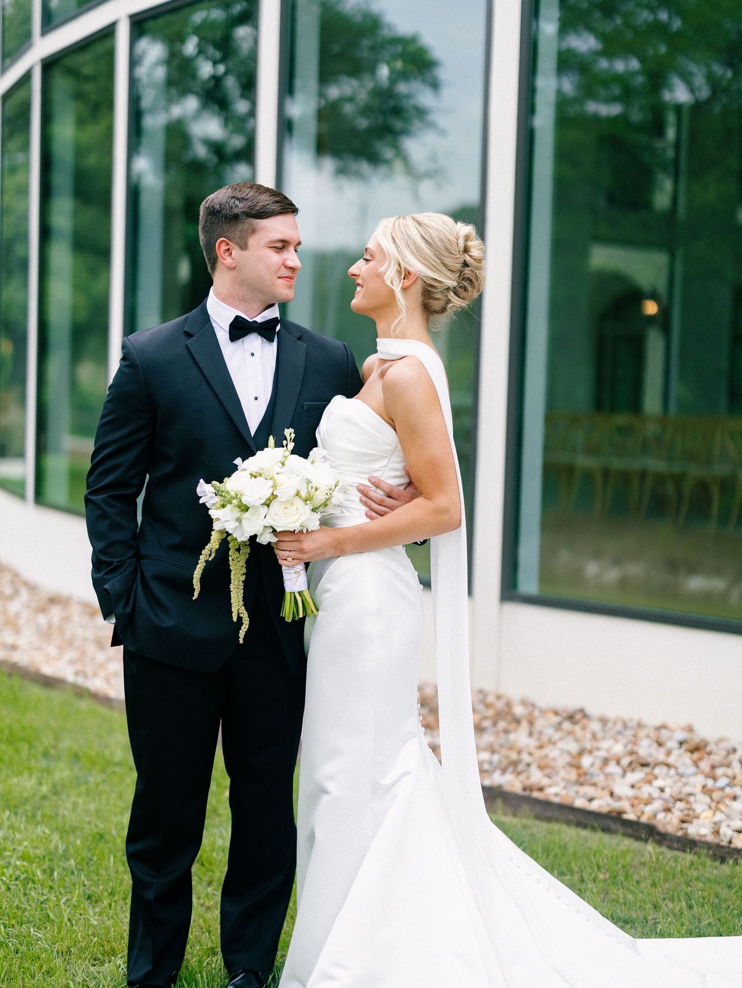 Bride in white gown holding bouquet stands with groom in black tuxedo outside glass building on wedding day.