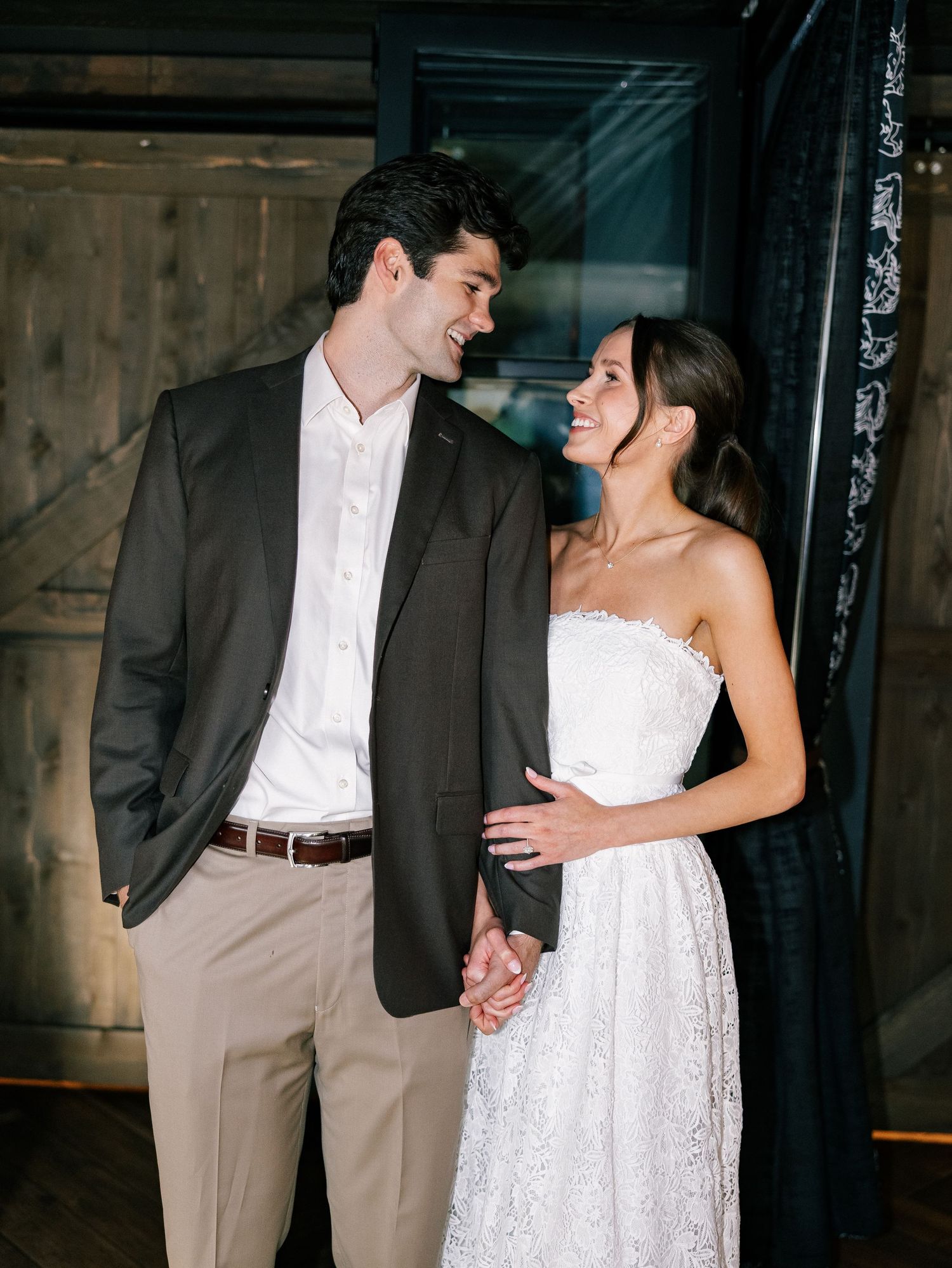 A bride in a strapless lace gown and groom in dark blazer share a romantic moment, smiling at each other.
