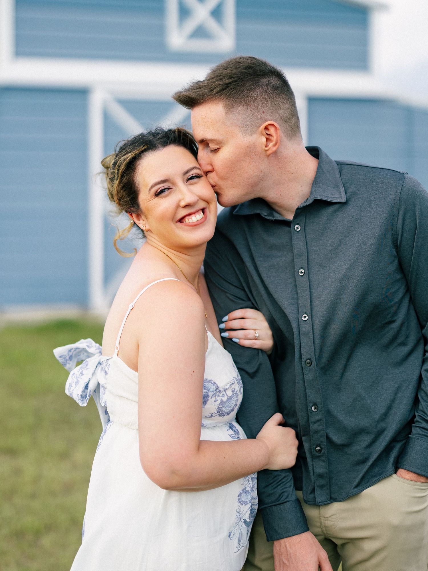 Couple posing outdoors near a blue barn, woman smiling as man kisses her cheek, engagement photo session.