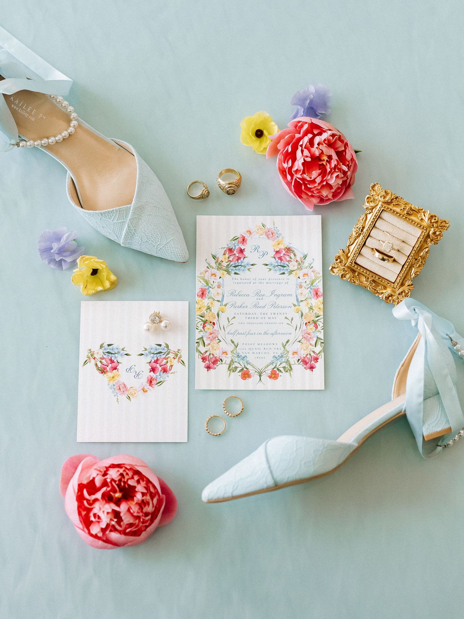 Elegant wedding flat lay with blue lace heels, floral invitation, pink peonies, gold rings, and pearl earrings on mint background.