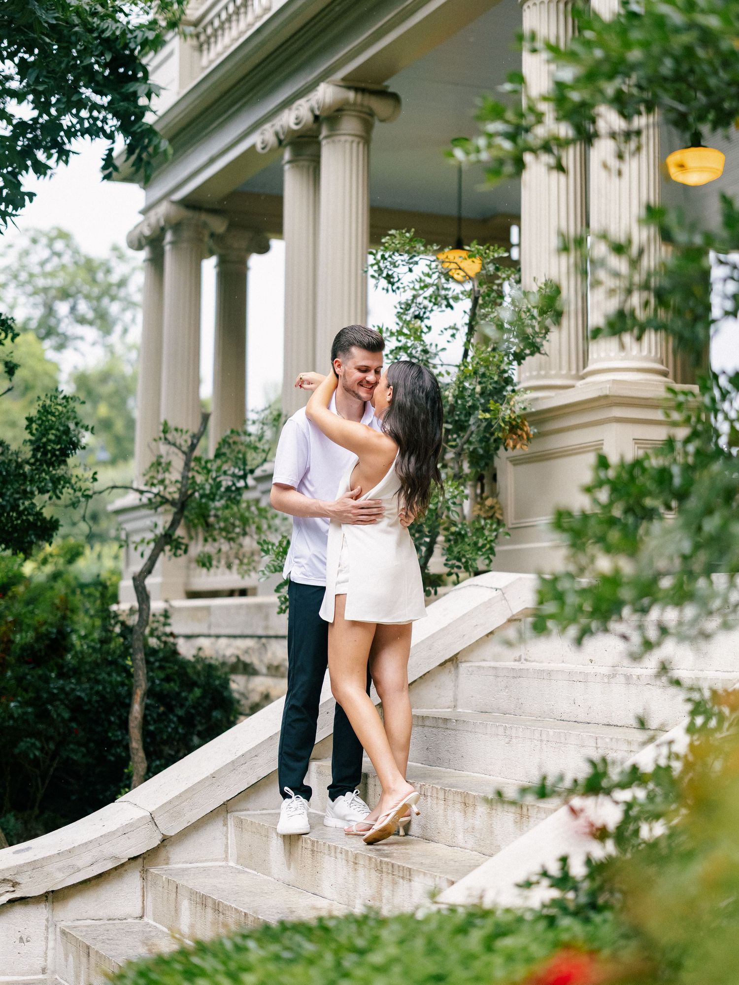Couple embracing and smiling on elegant stone steps of a classical columned mansion surrounded by lush greenery.