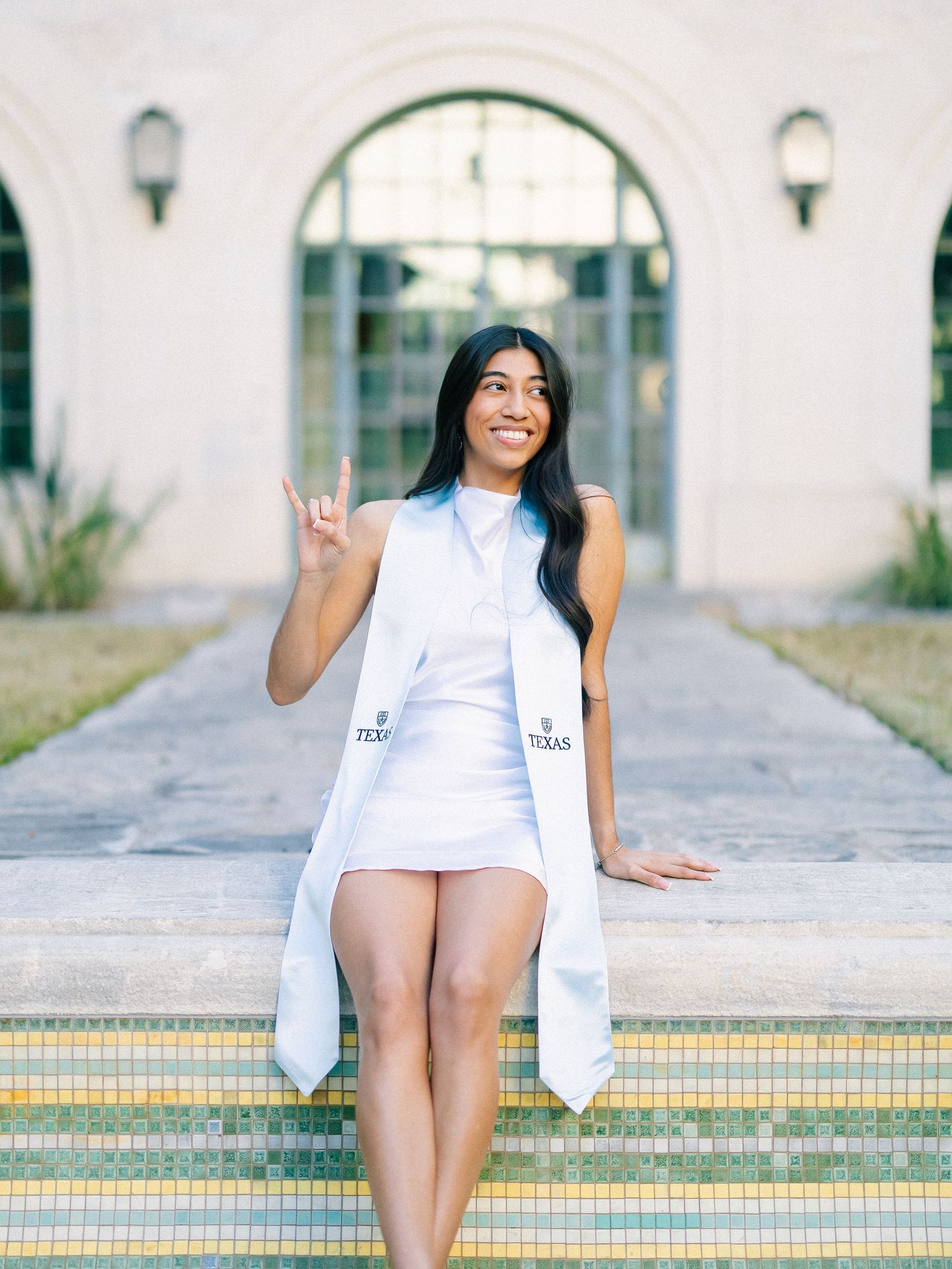 A young woman in a white dress and graduation stole poses outdoors, flashing a hand sign, celebrating her Texas graduation.