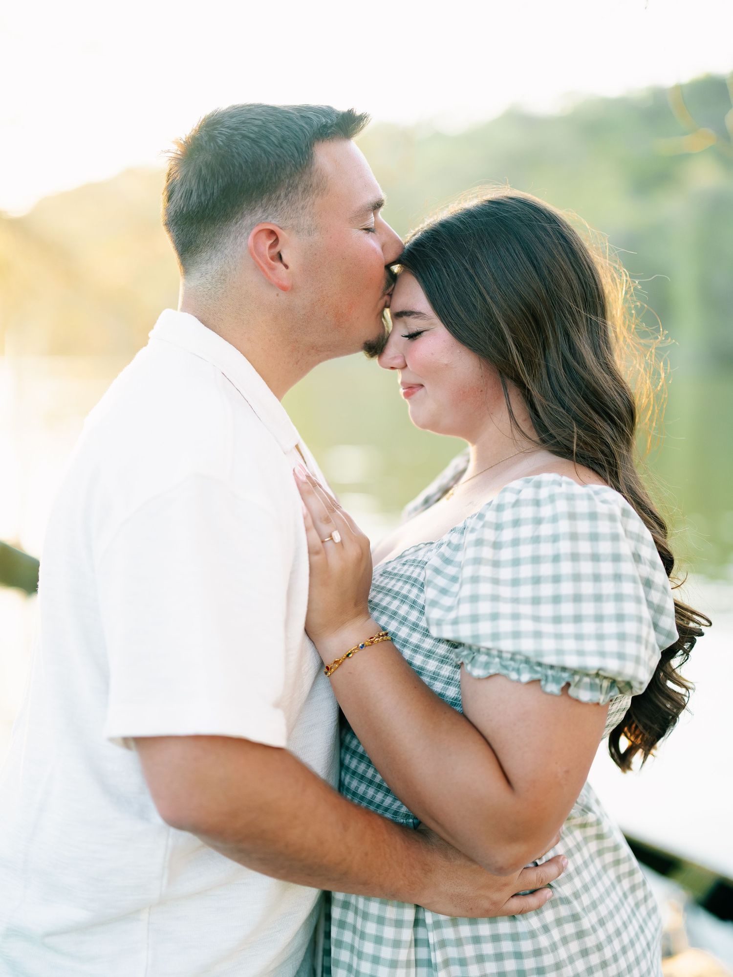 Romantic couple sharing a tender forehead kiss by a shimmering lake at golden hour.