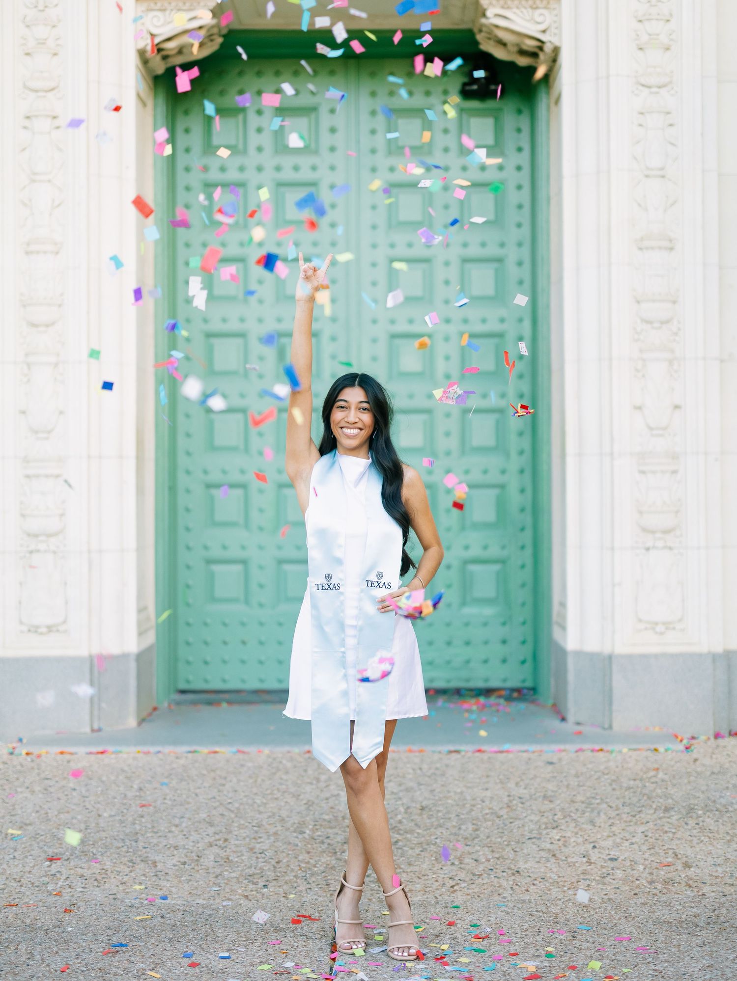 Joyful graduate in white dress celebrates with colorful confetti in front of green doors.