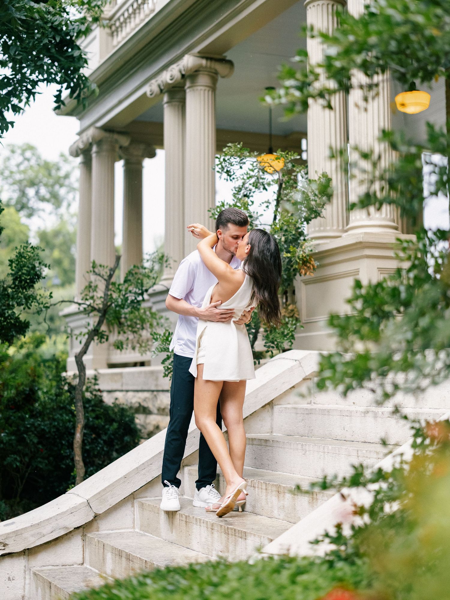 Couple sharing a romantic kiss on stone steps outside a classical building with tall columns and lush greenery.