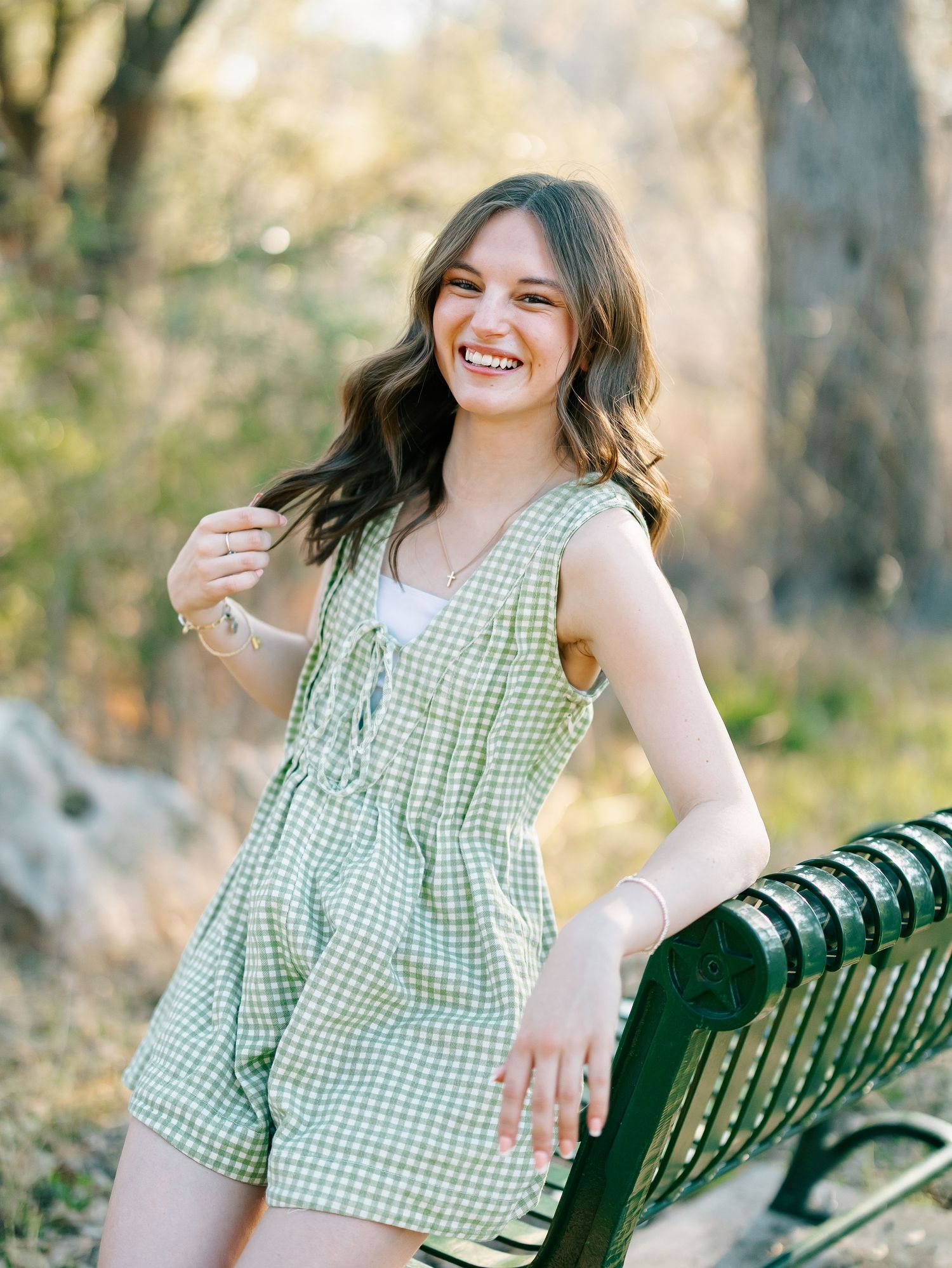 A smiling young woman in a green gingham romper leans on a park bench outdoors.
