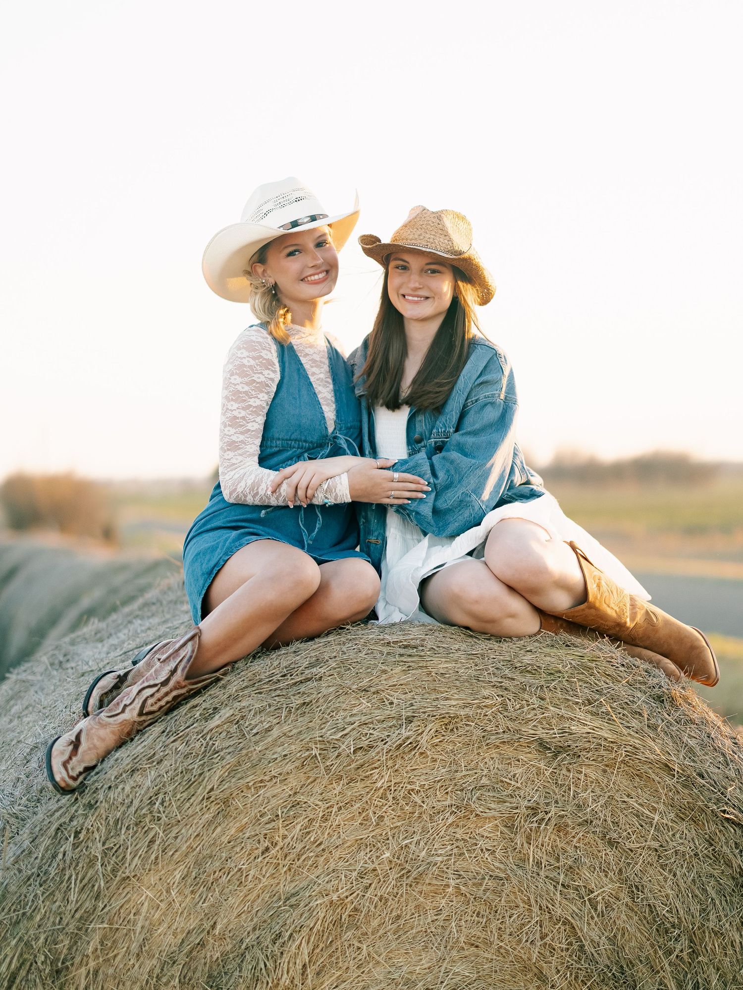 Two women in cowboy hats and denim outfits sit smiling on a hay bale in a sunny field.