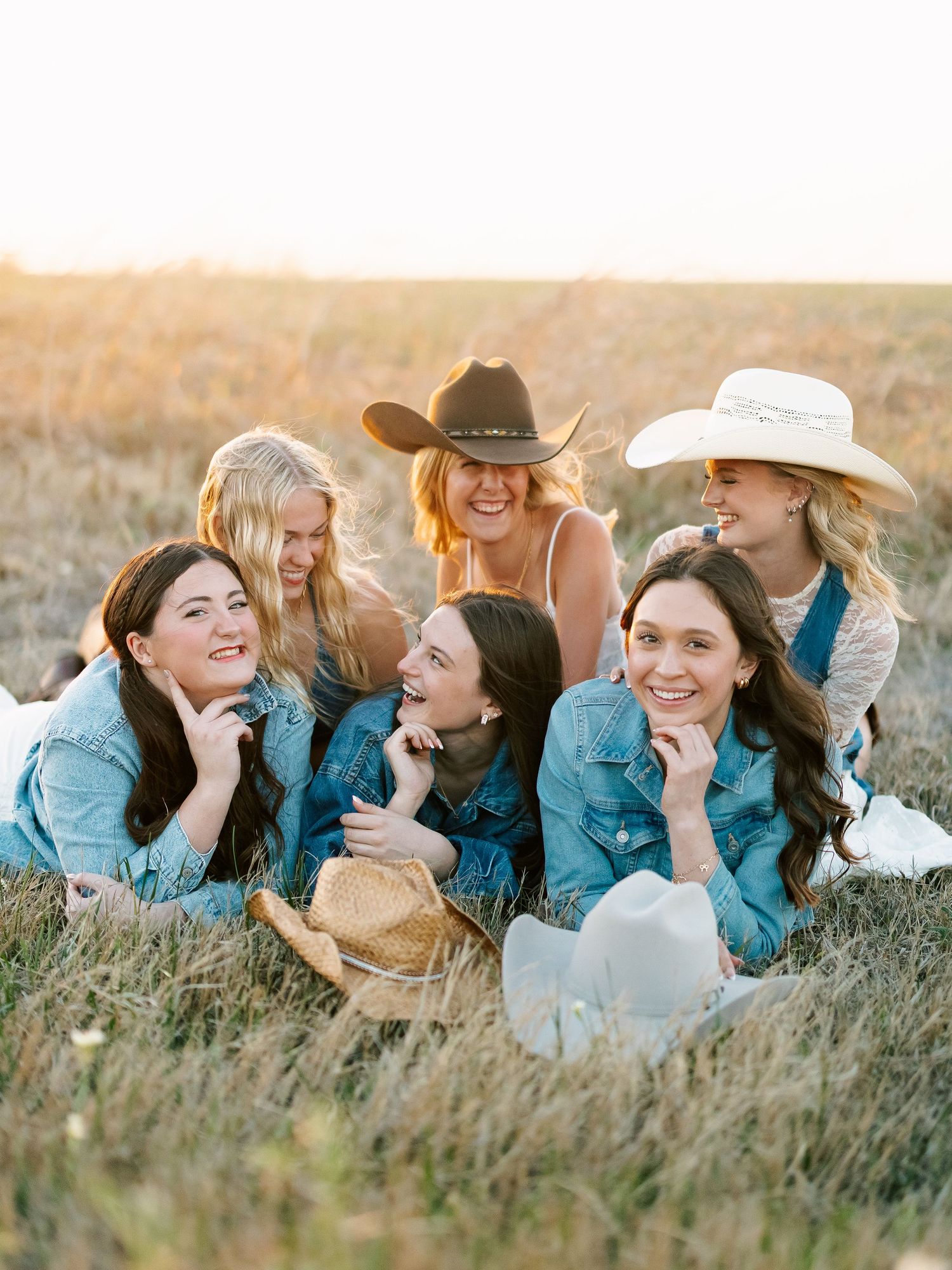 Six women in denim and cowboy hats laughing together in a golden field at sunset.