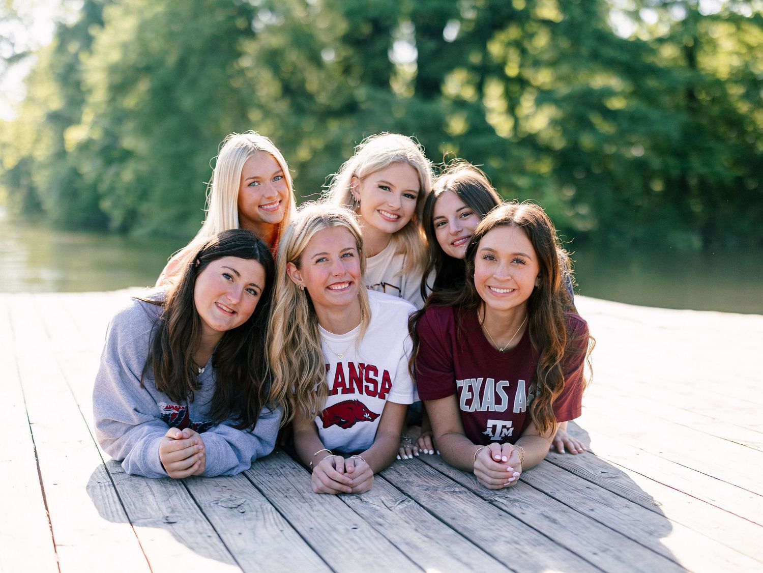 Six smiling young women in college sweatshirts pose together on a wooden dock near a lake surrounded by trees.