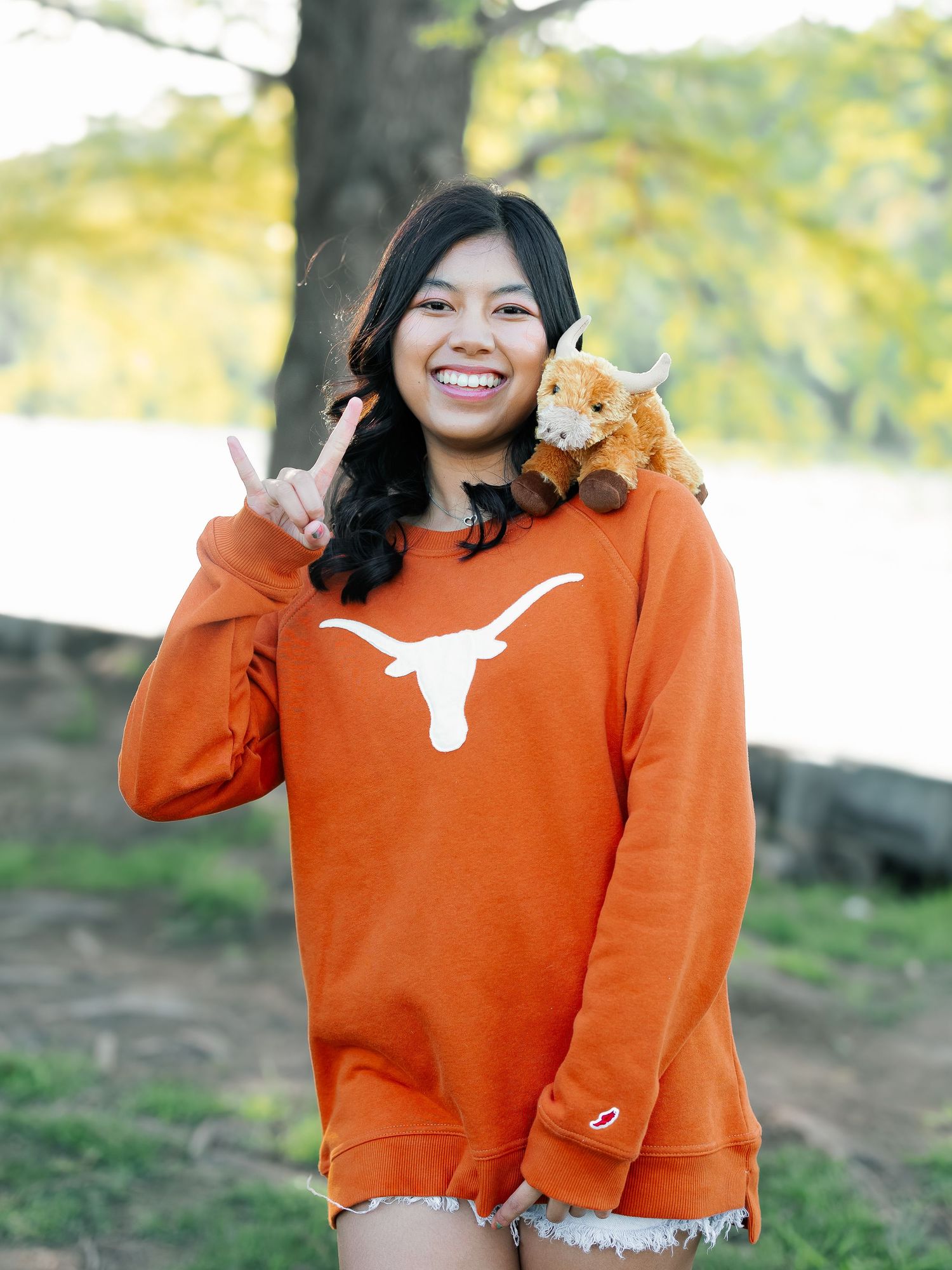 Smiling young woman in orange Texas Longhorns sweatshirt making hook em sign with stuffed longhorn on her shoulder.