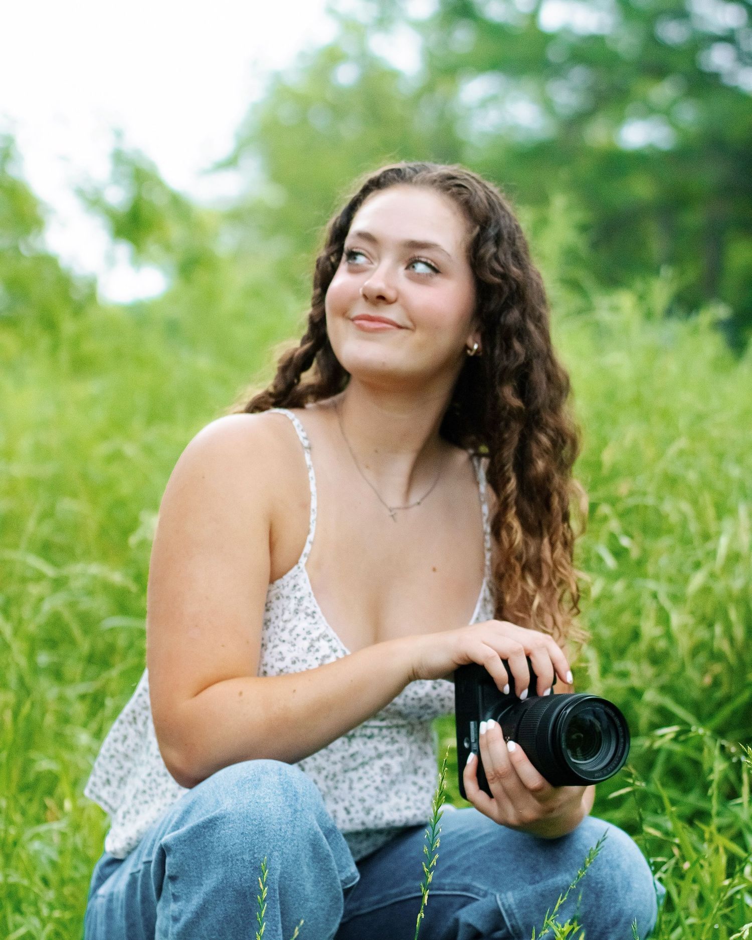 Young woman with curly hair holding a camera while sitting in a lush green field, looking up thoughtfully.