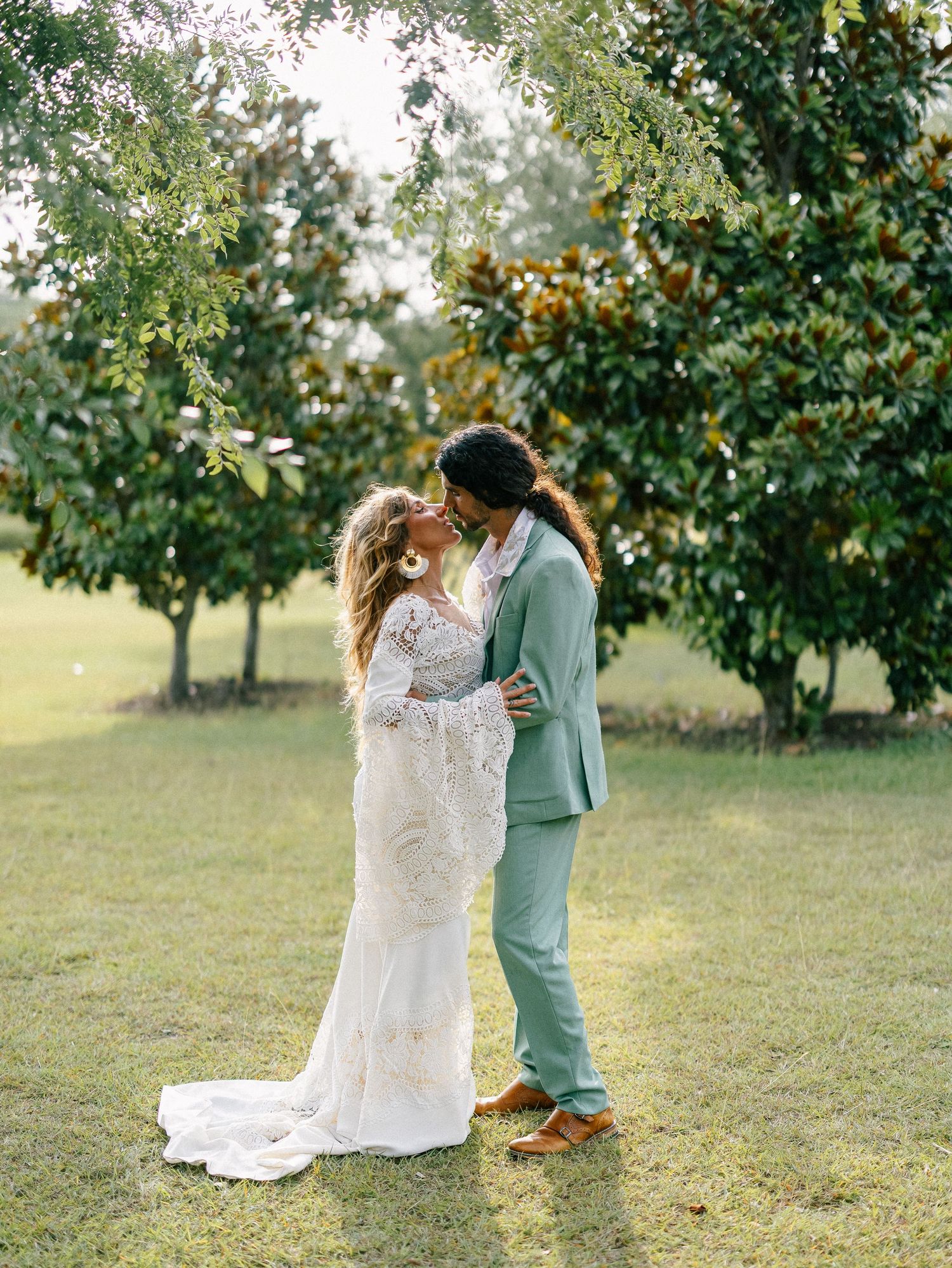 A bride and groom share a kiss outdoors on green grass, surrounded by lush trees, she in lace gown, he in sage suit.