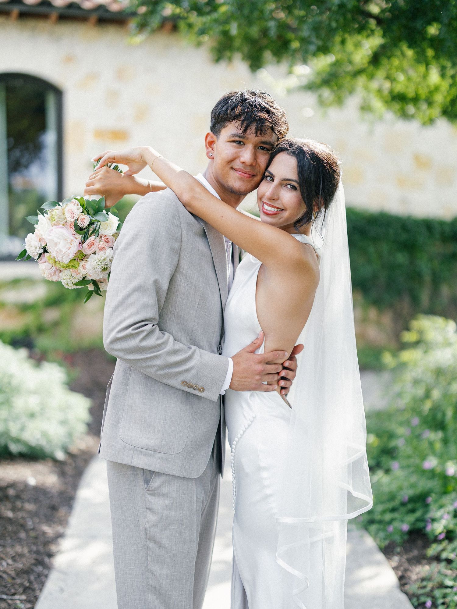 A bride and groom pose outdoors, she holds pink bouquet and wears veil, he wears light gray suit.