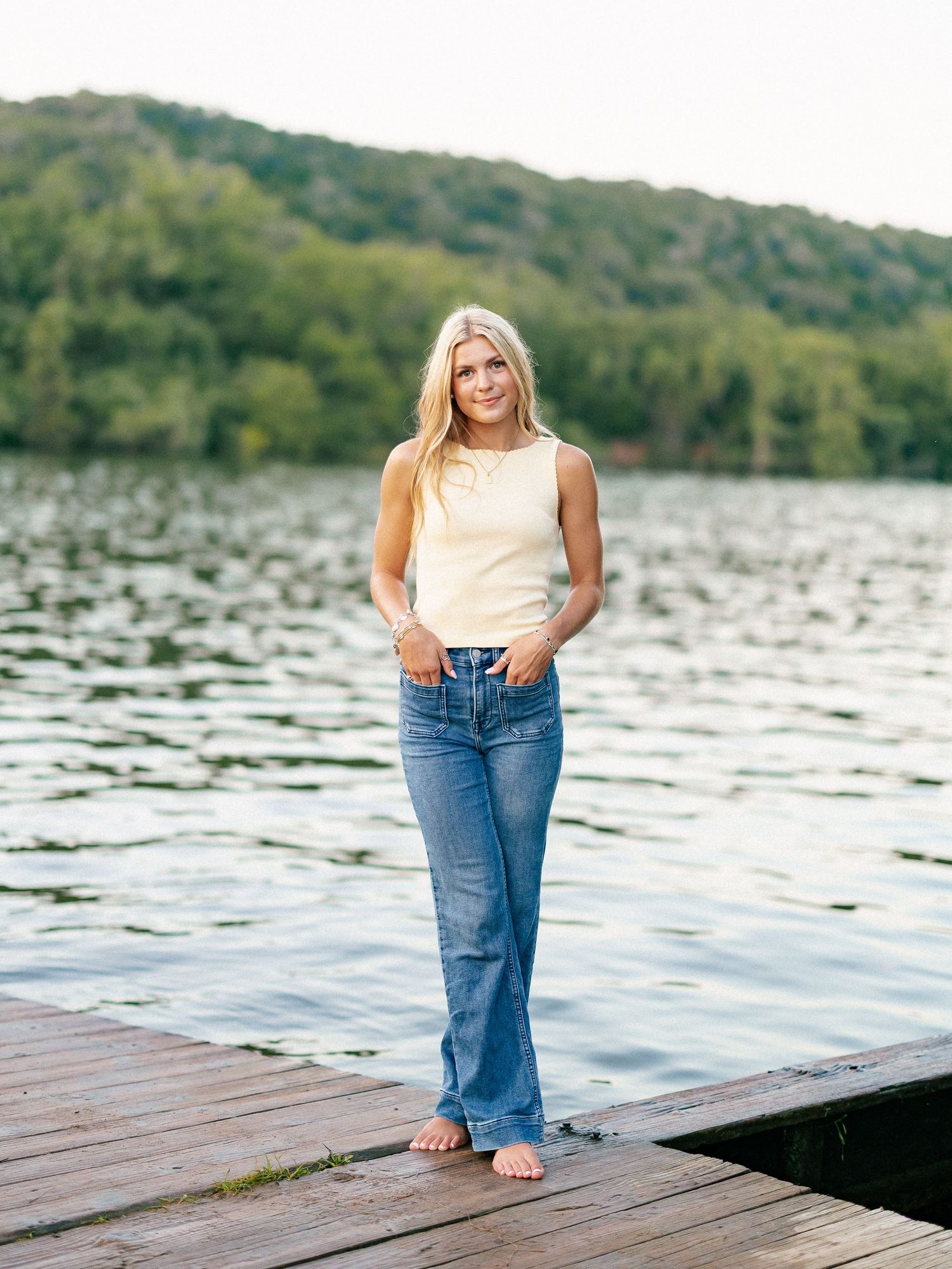 Blonde woman in cream top and flare jeans standing barefoot on a wooden dock by a serene lake.