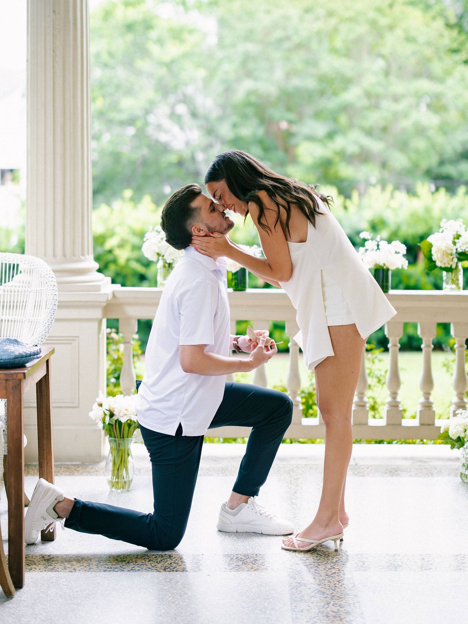 Man on one knee proposing to woman on a elegant porch, both leaning in for a kiss during the proposal.