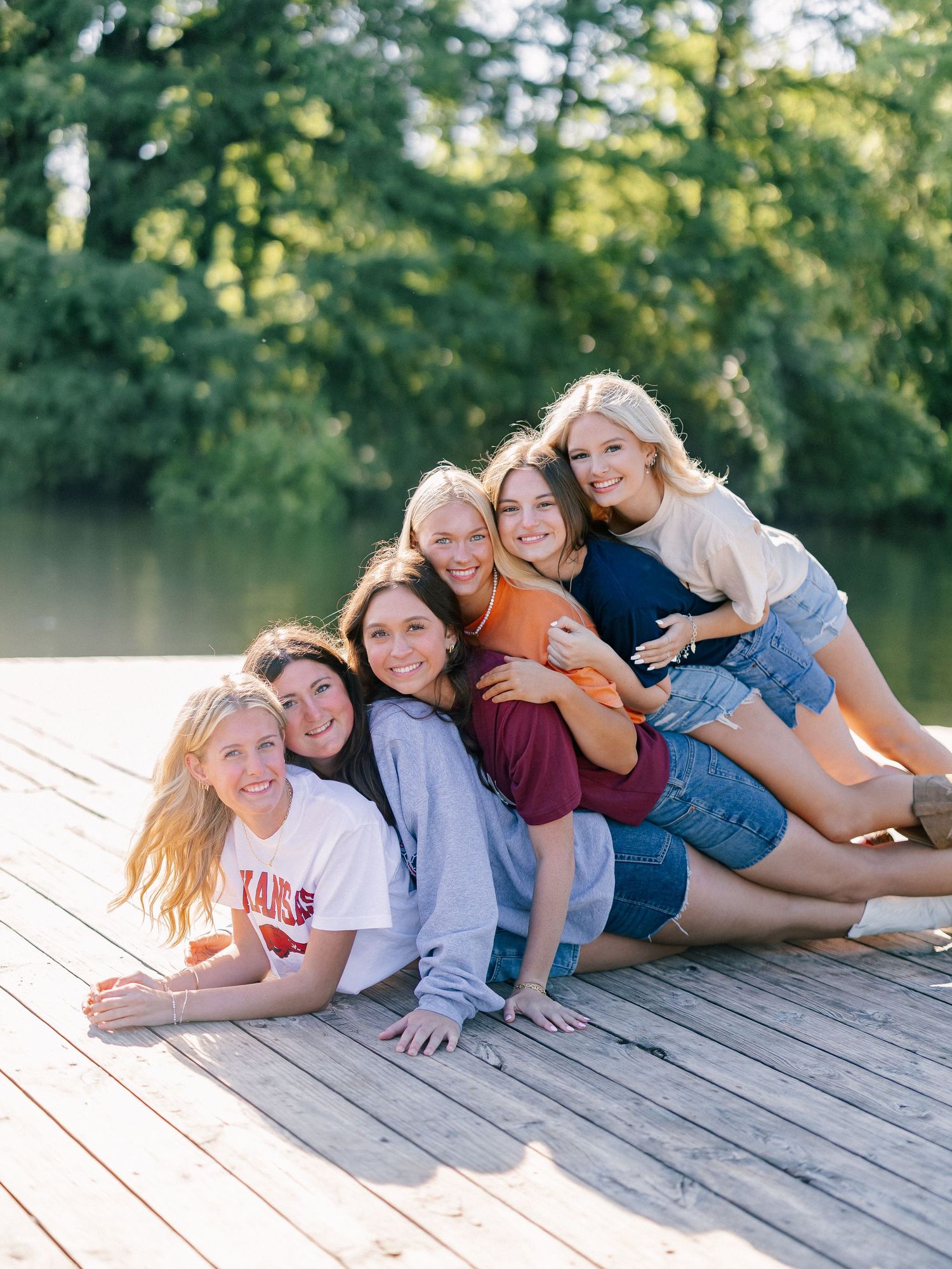 Six young women pose together laughing on a wooden dock by a lake surrounded by lush green trees on a sunny day.