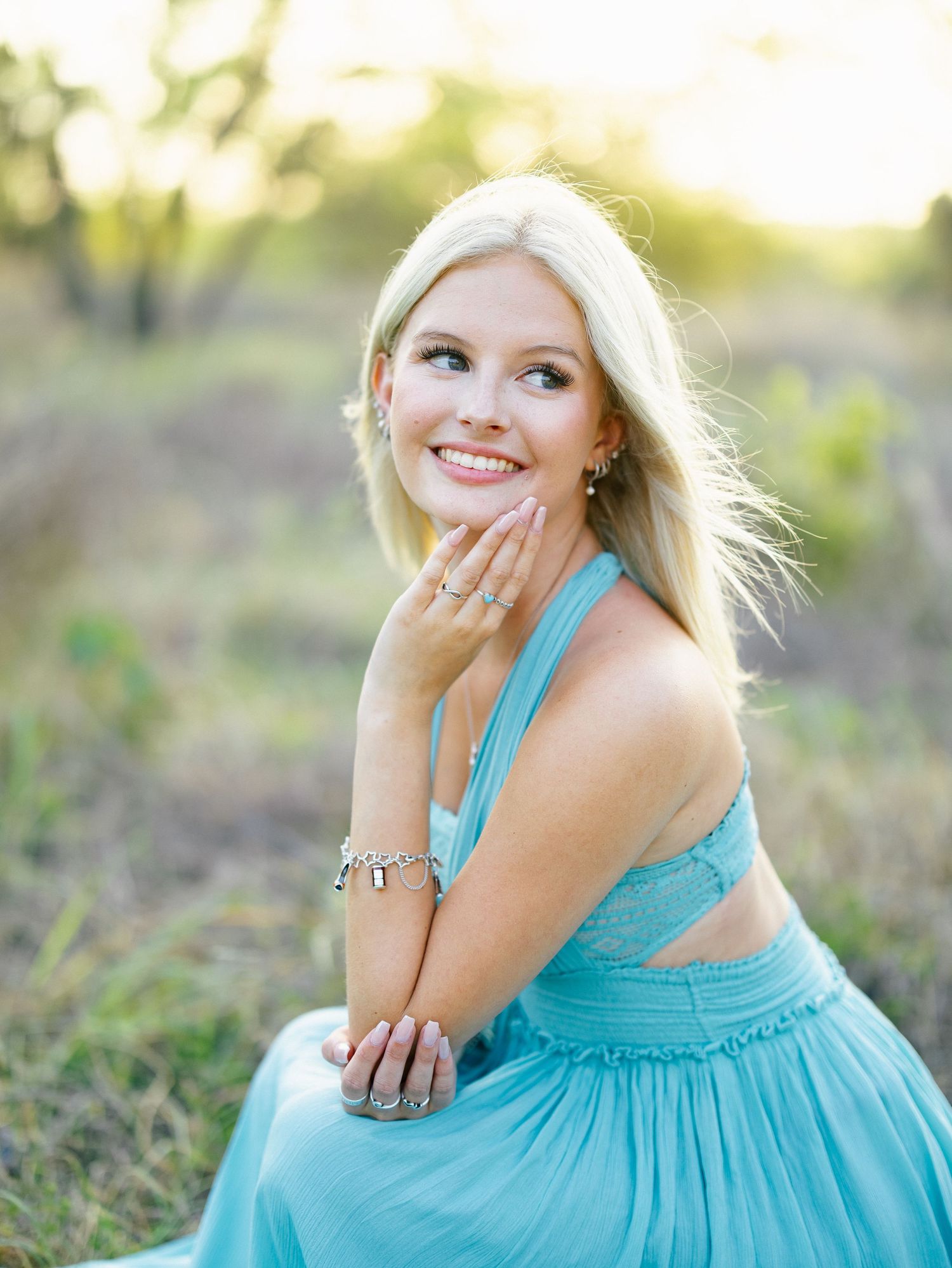 Smiling blonde woman in teal halter dress posing outdoors wearing jewelry in golden hour light.
