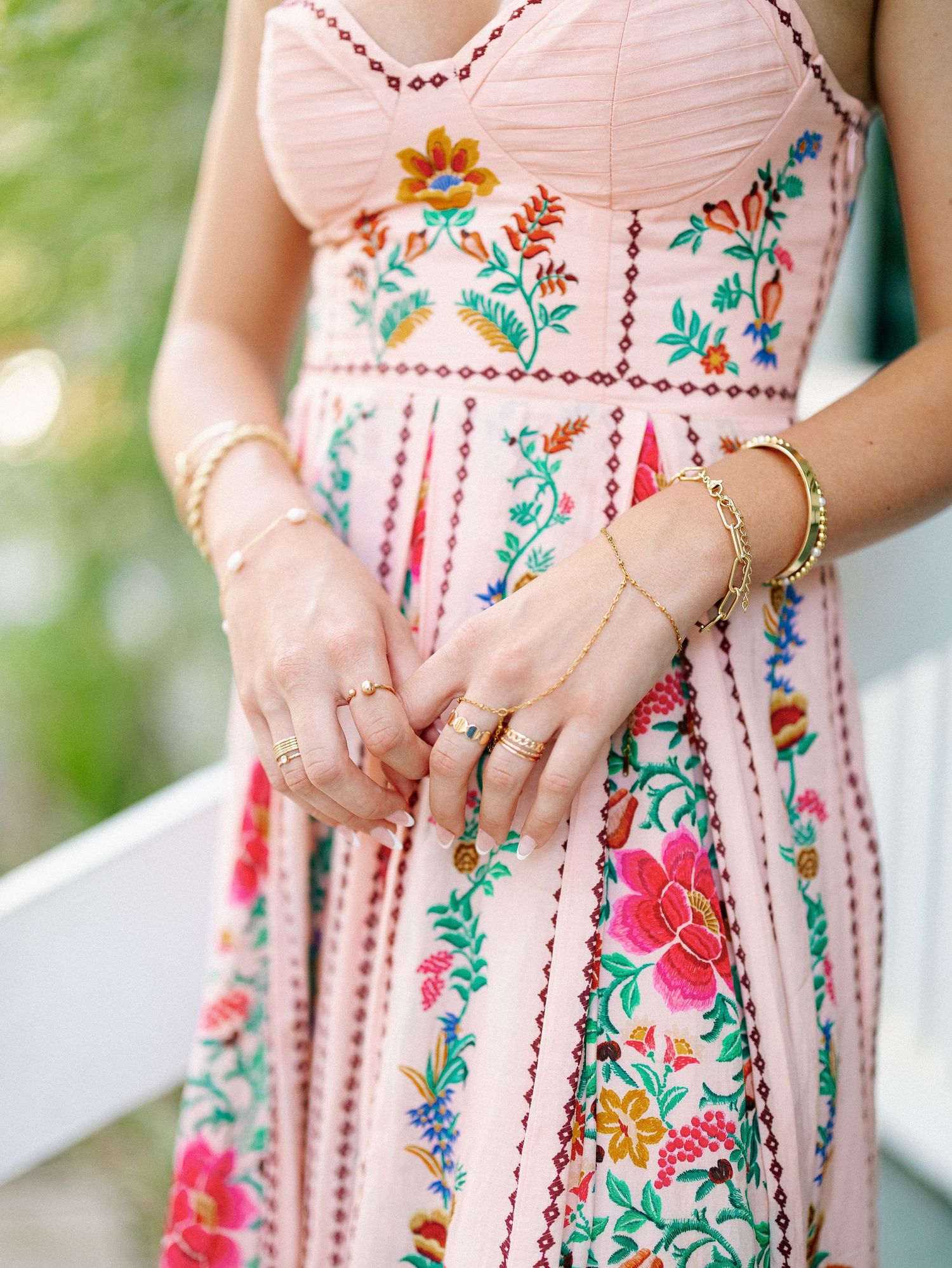Woman wearing a pink floral embroidered maxi dress with gold hand chain bracelet and rings.