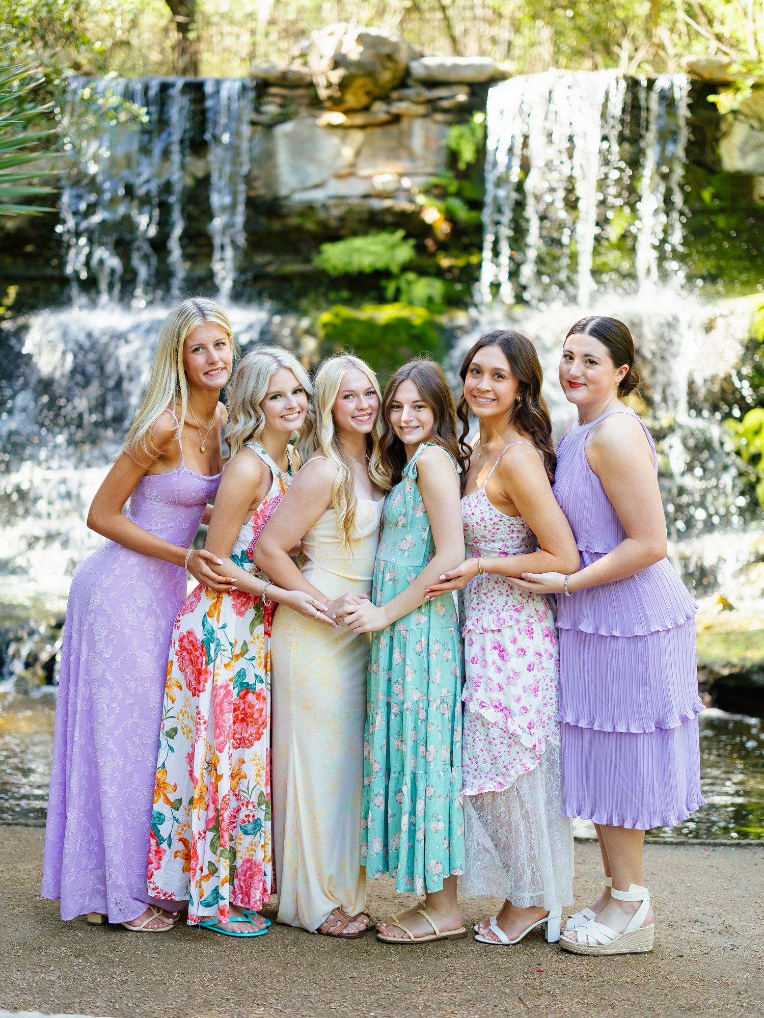 Seven young women in pastel dresses pose together smiling in front of a beautiful waterfall outdoors.