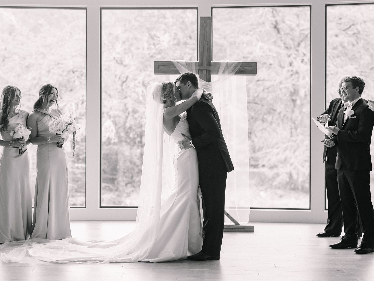 Black and white wedding photo of couple sharing first kiss at altar with cross, bridesmaids, and officiant in chapel.