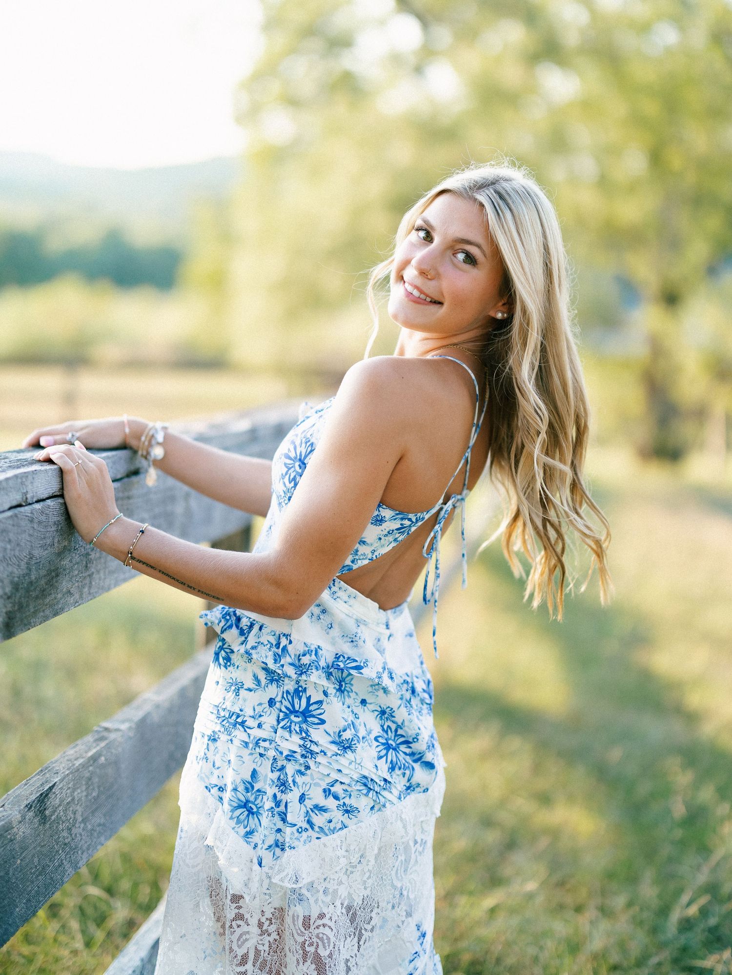 Young woman in blue floral lace dress leaning on rustic wooden fence in sunny green meadow.
