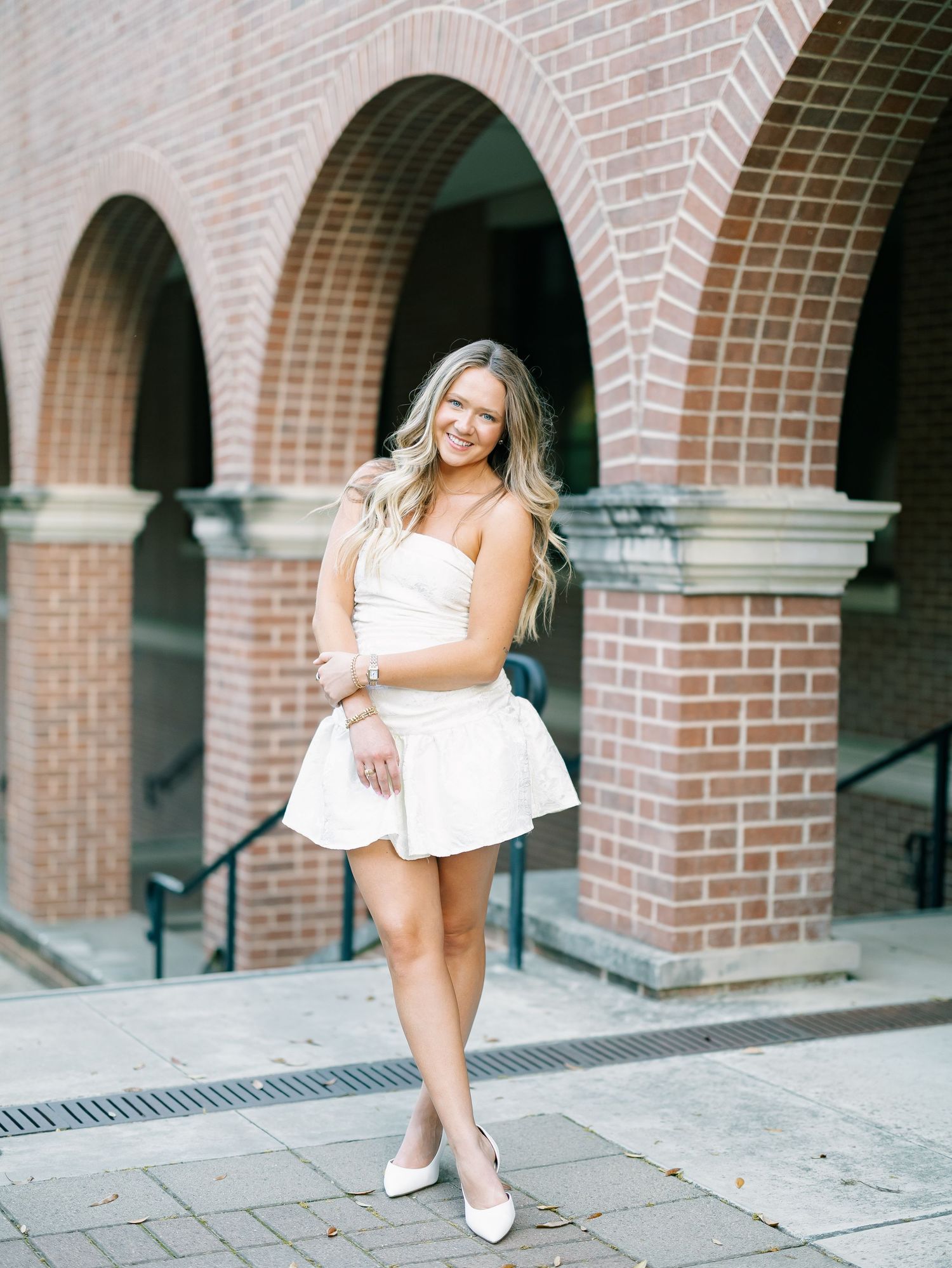 Smiling blonde woman in white strapless mini dress posing under brick arches outdoors.