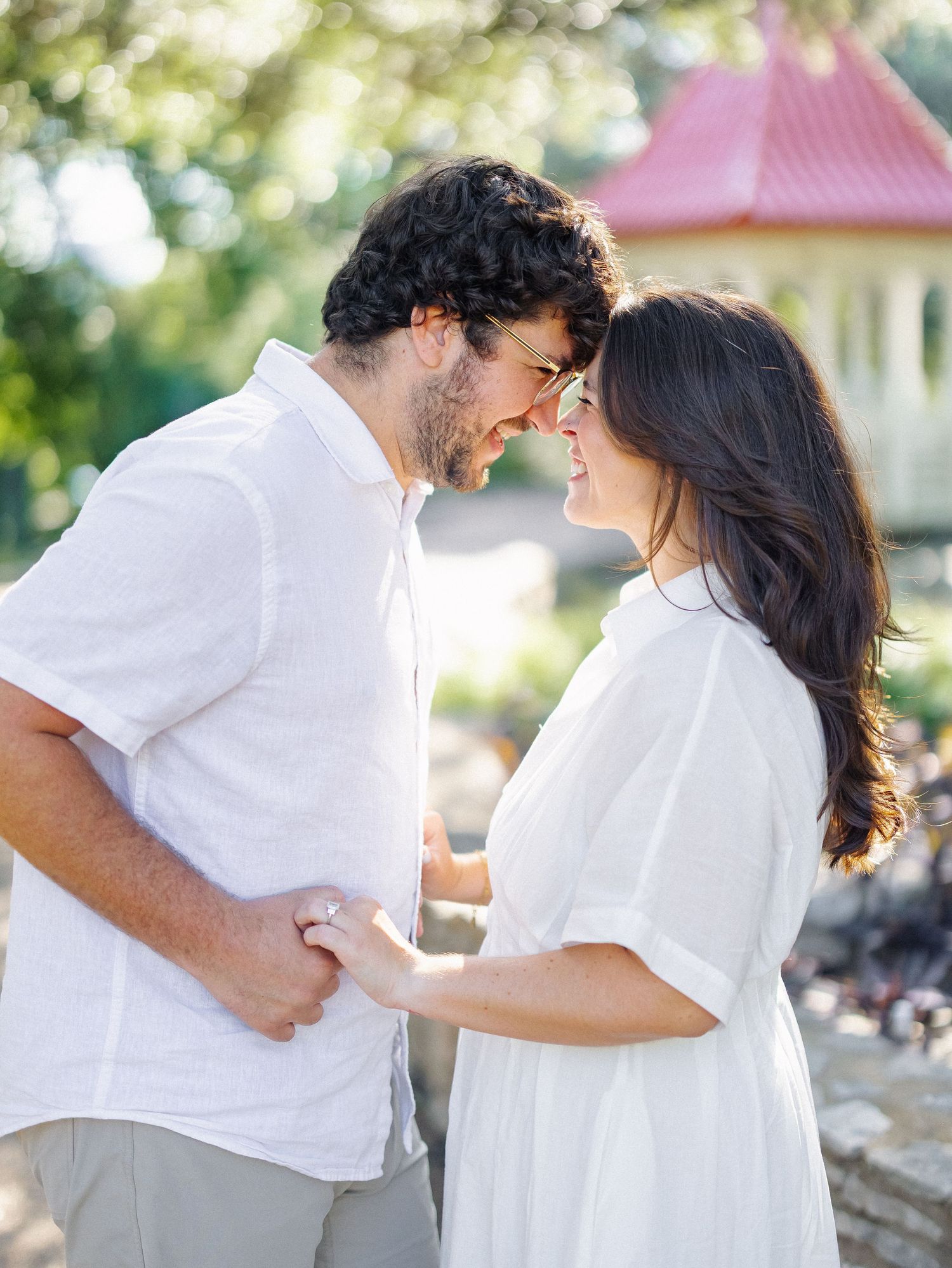 Happy couple in matching white outfits smiling at each other outdoors near a charming red gazebo.
