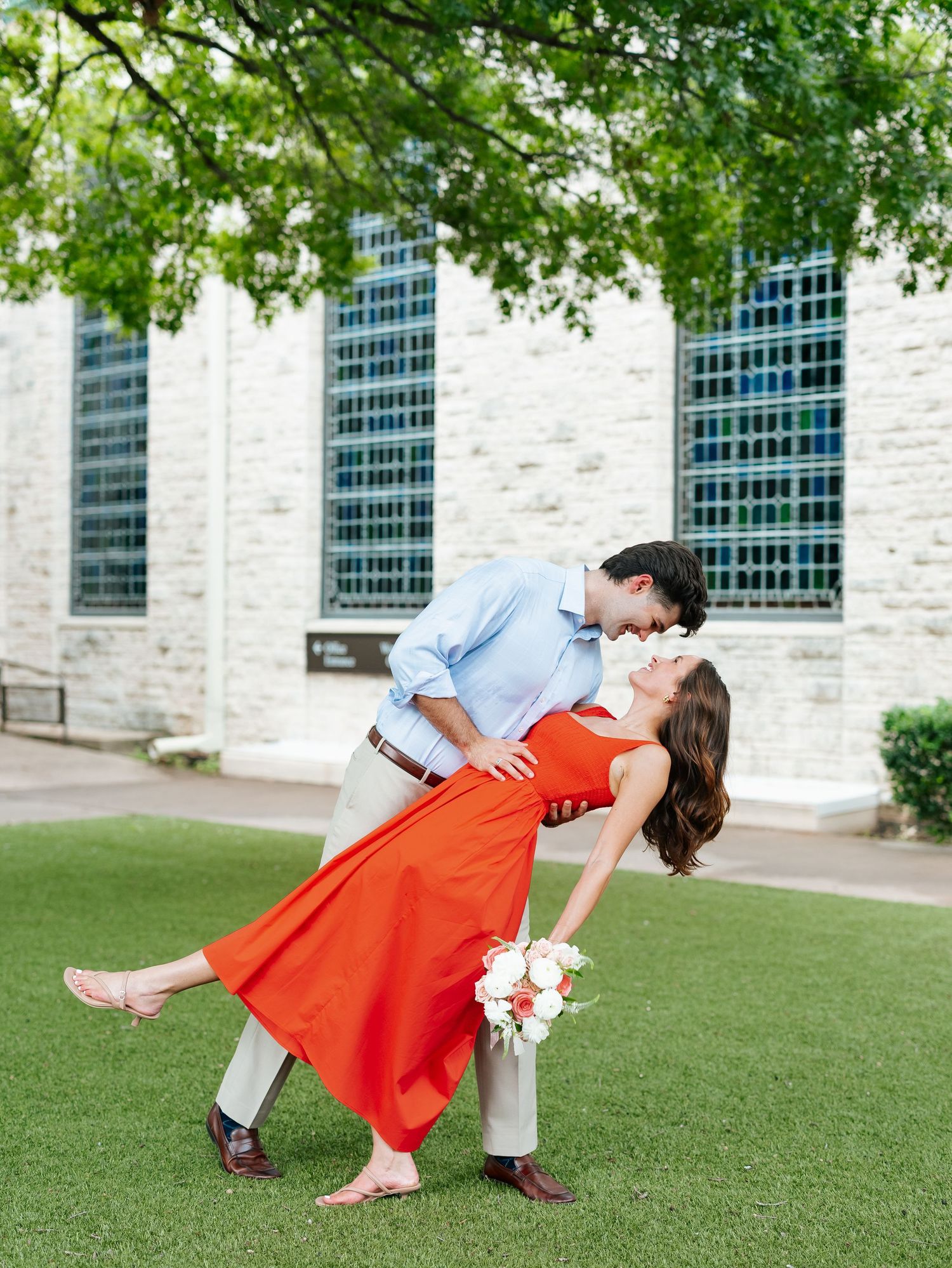 Man dipping woman in red dress holding white flowers while kissing outdoors on green lawn.