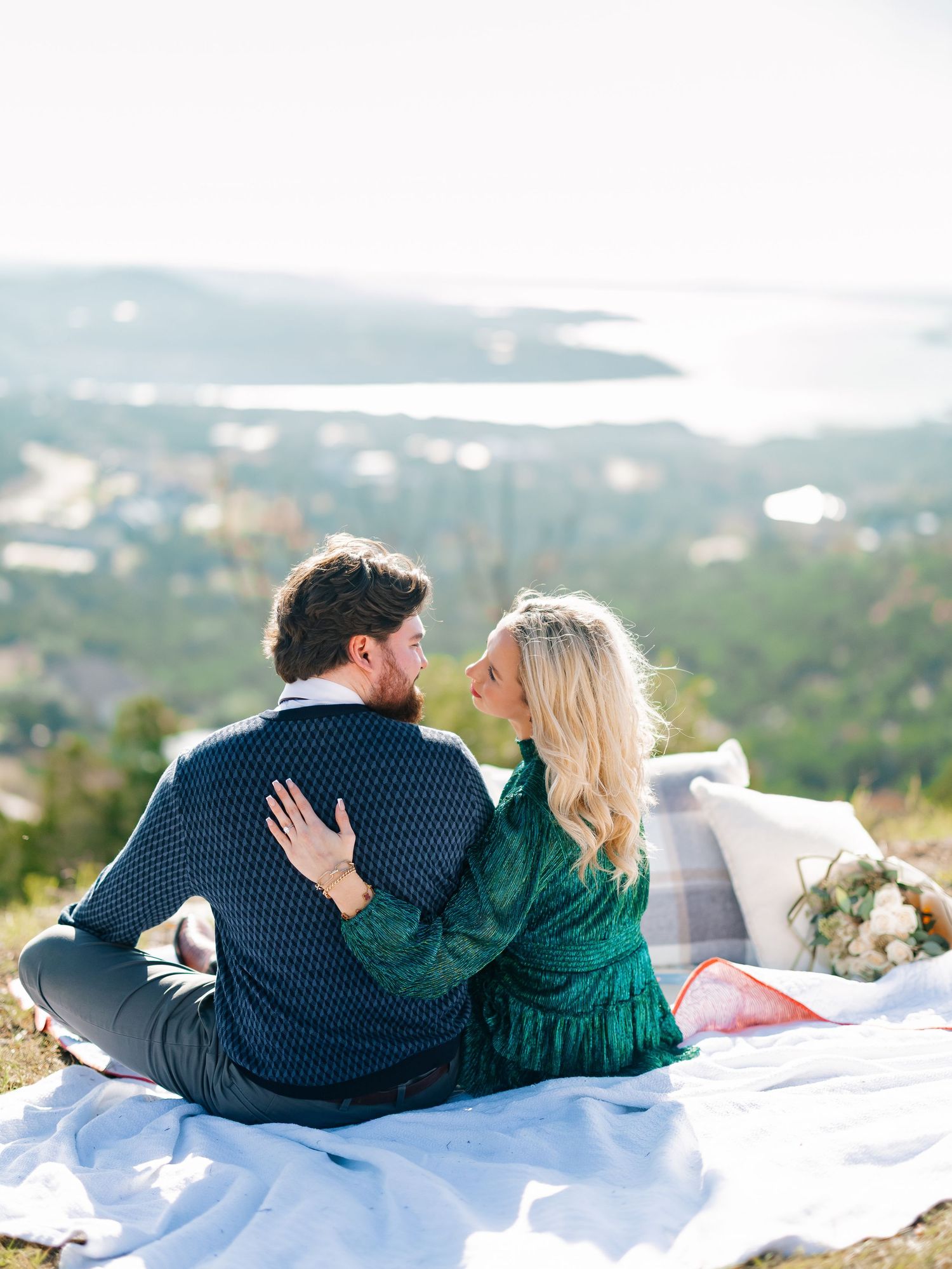 Couple sitting closely on a blanket outdoors, gazing at each other with a scenic view.