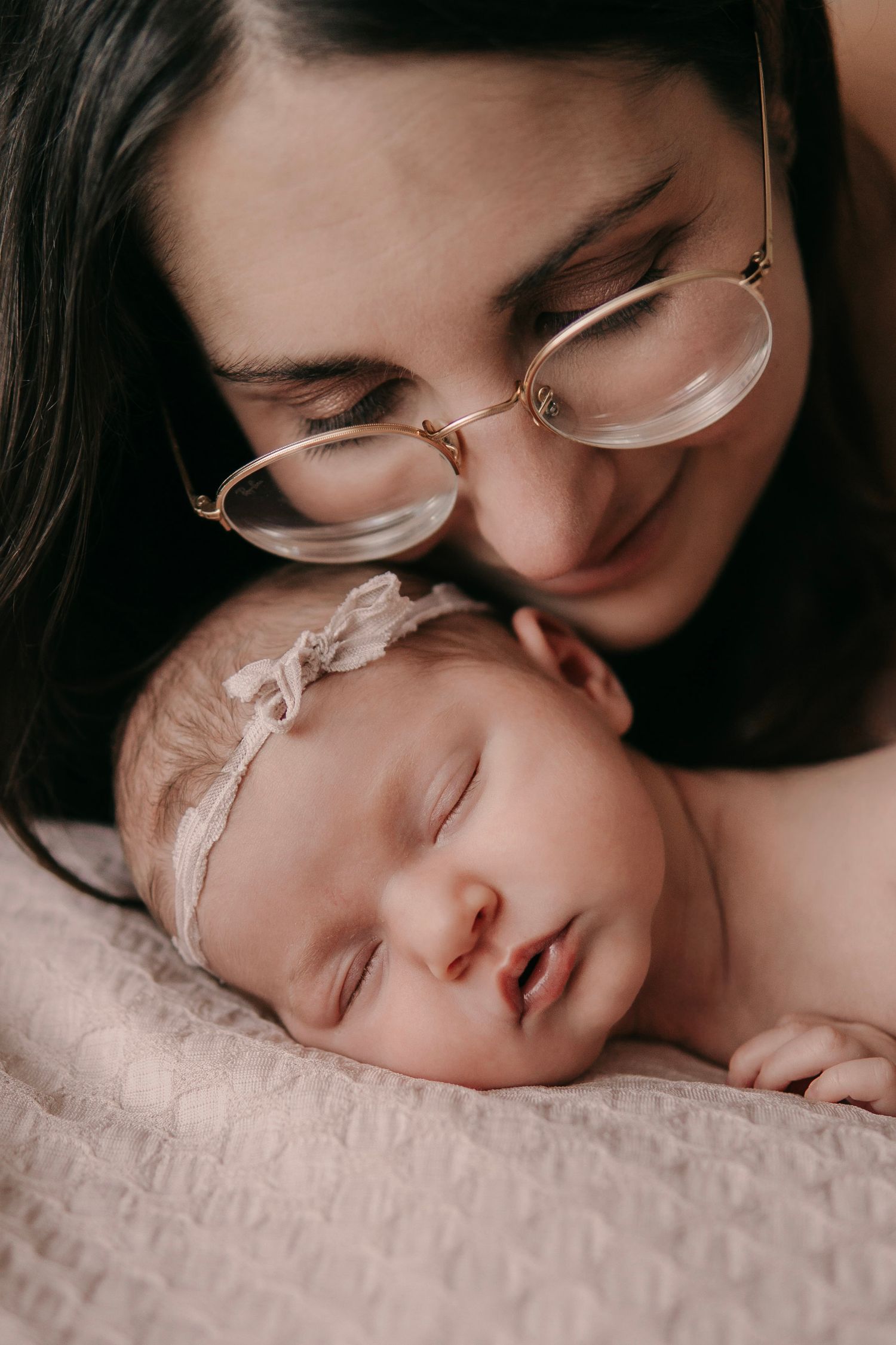 Mother with glasses gently kissing her sleeping newborn baby girl.