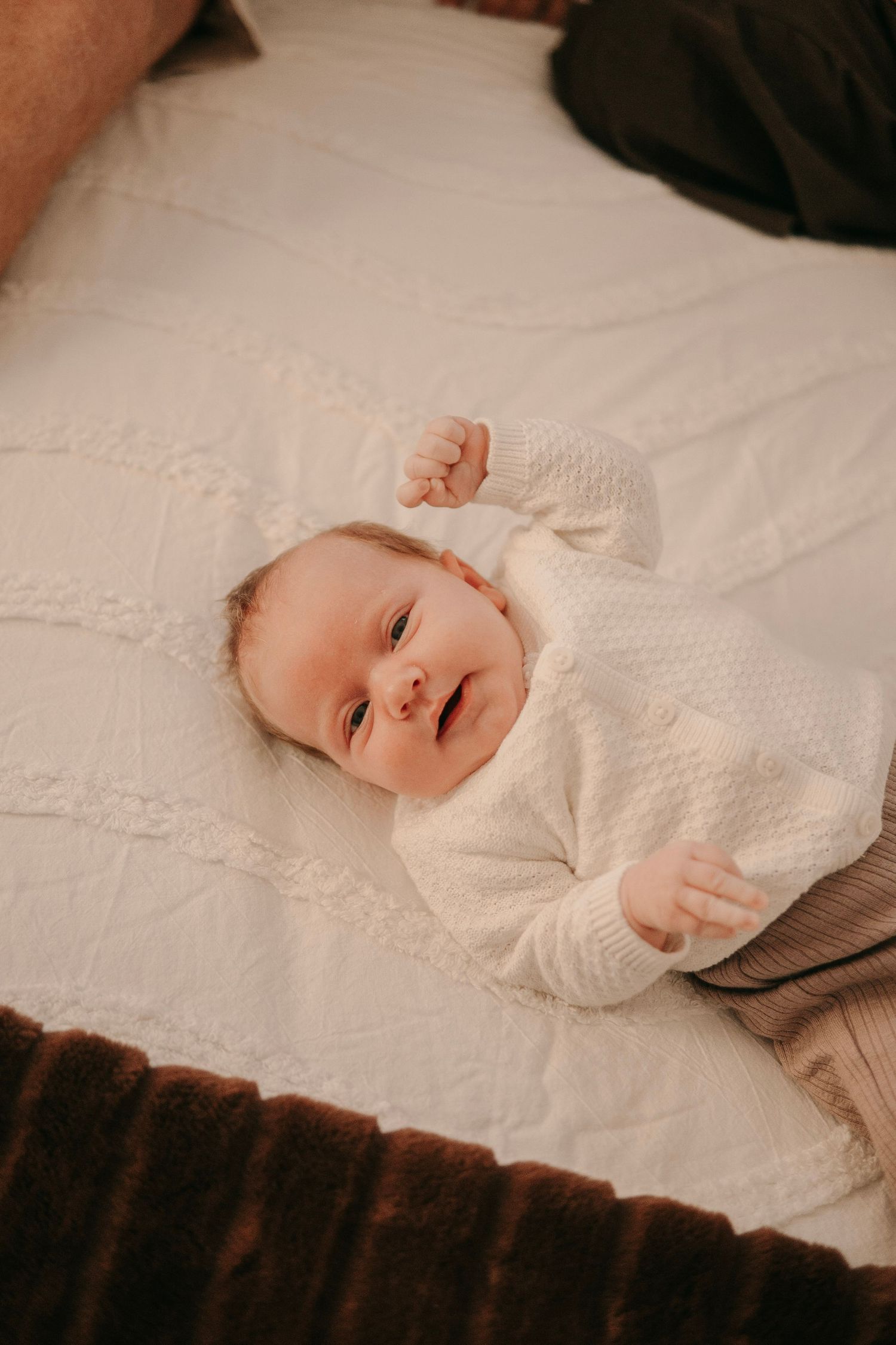 A smiling baby in a white knit sweater lying on a white textured blanket.