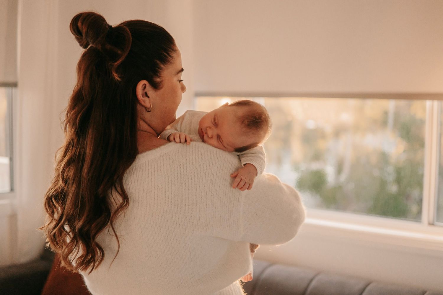 Young mother in white sweater gently holding a sleeping newborn baby against her shoulder indoors.