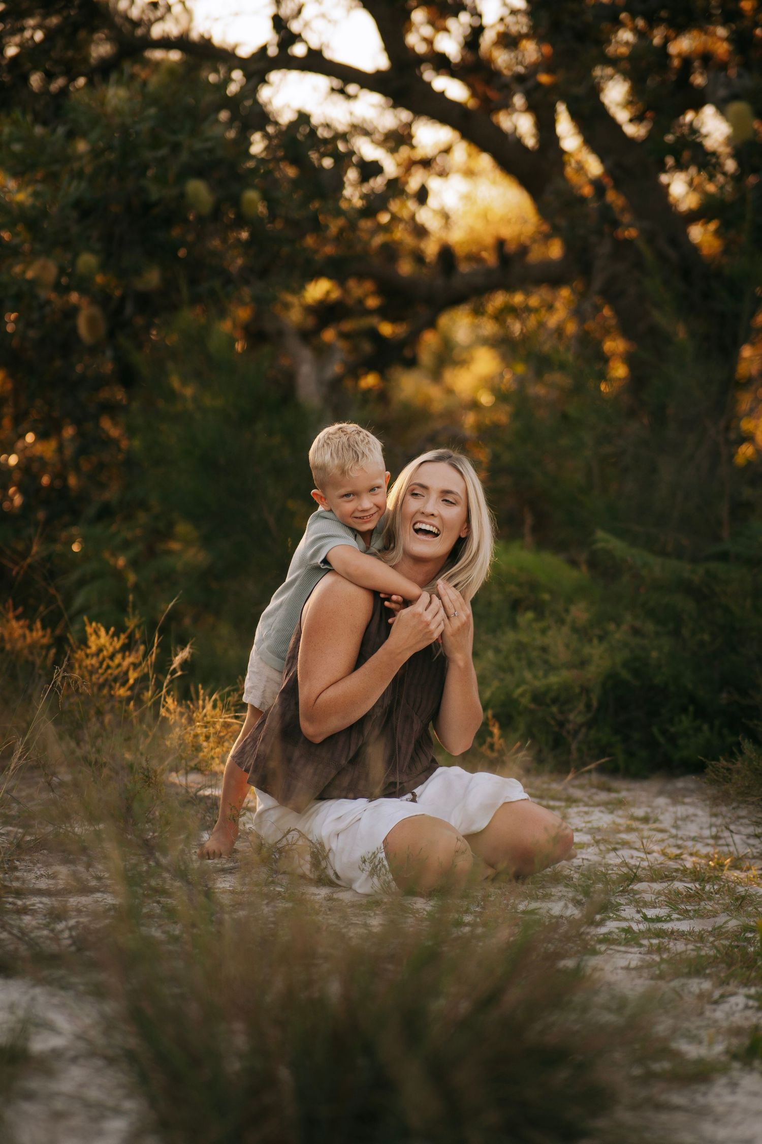 A laughing blonde mother being hugged from behind by her young son outdoors.
