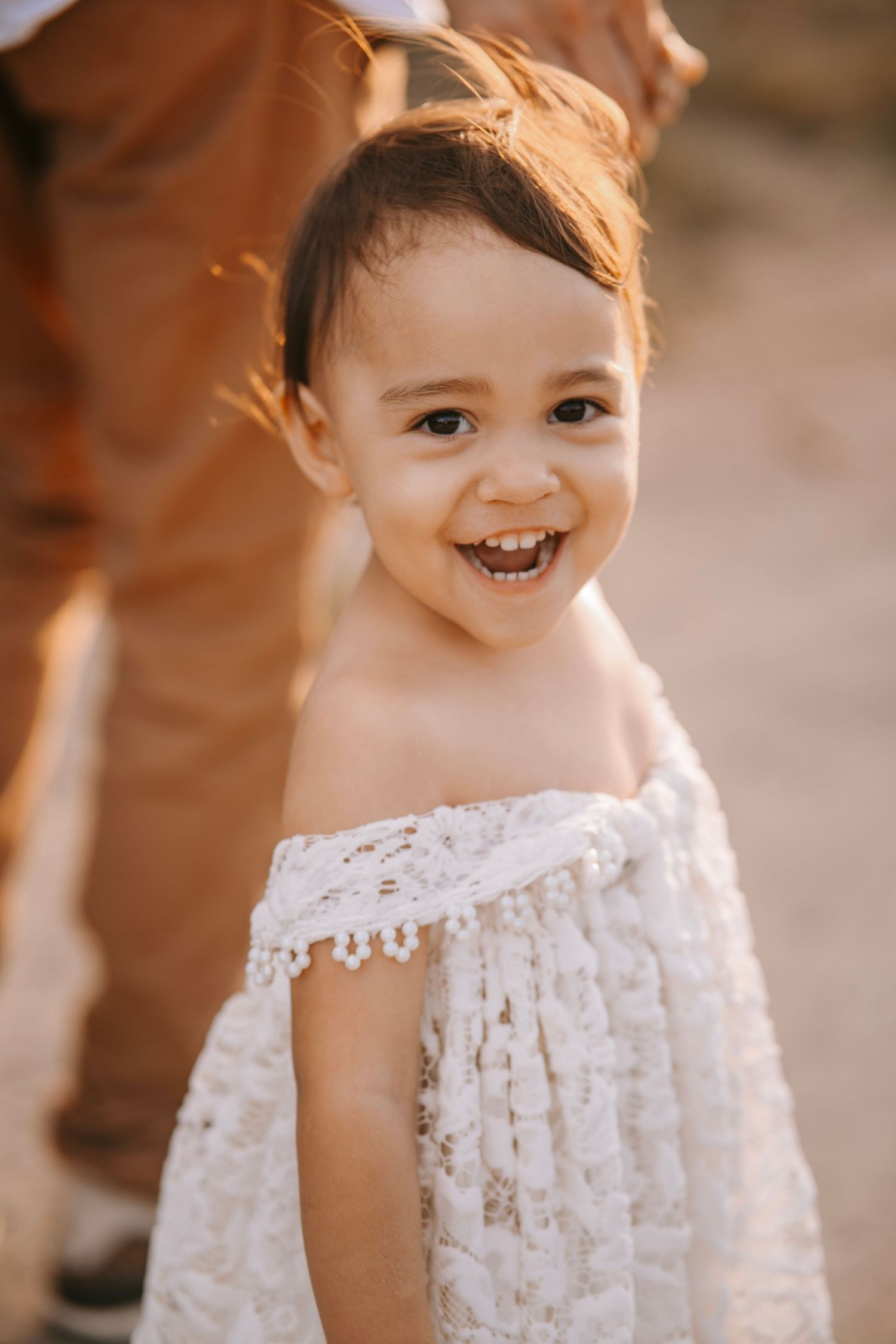 Adorable smiling toddler girl in white lace off-shoulder dress laughing in warm sunlight.