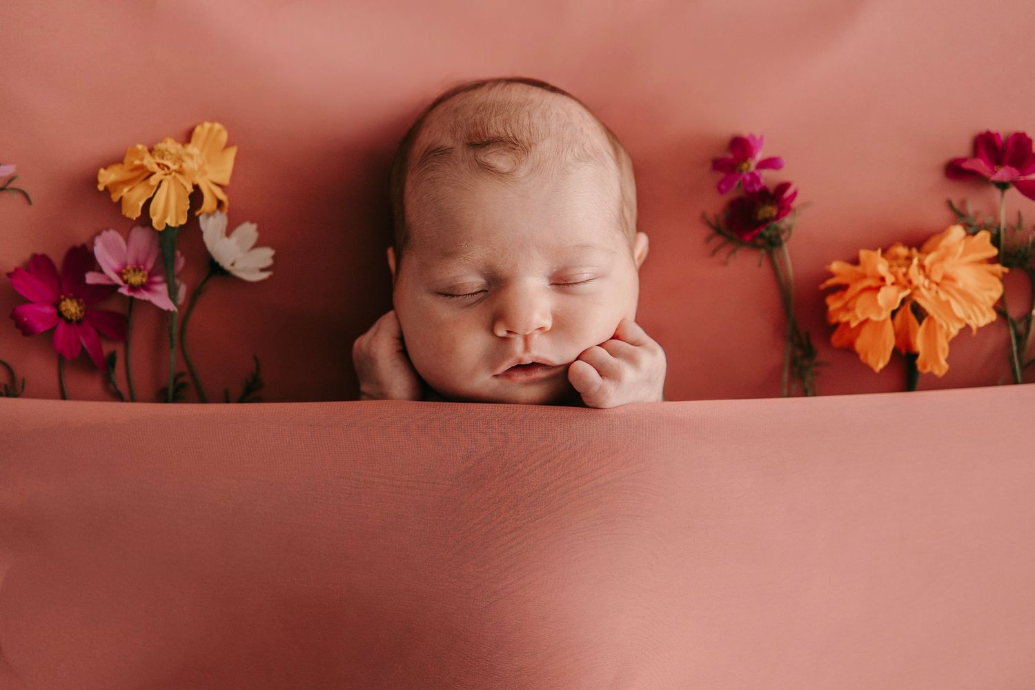 Sleeping newborn baby posed with colorful pink, orange, and white flowers on a soft coral pink backdrop.