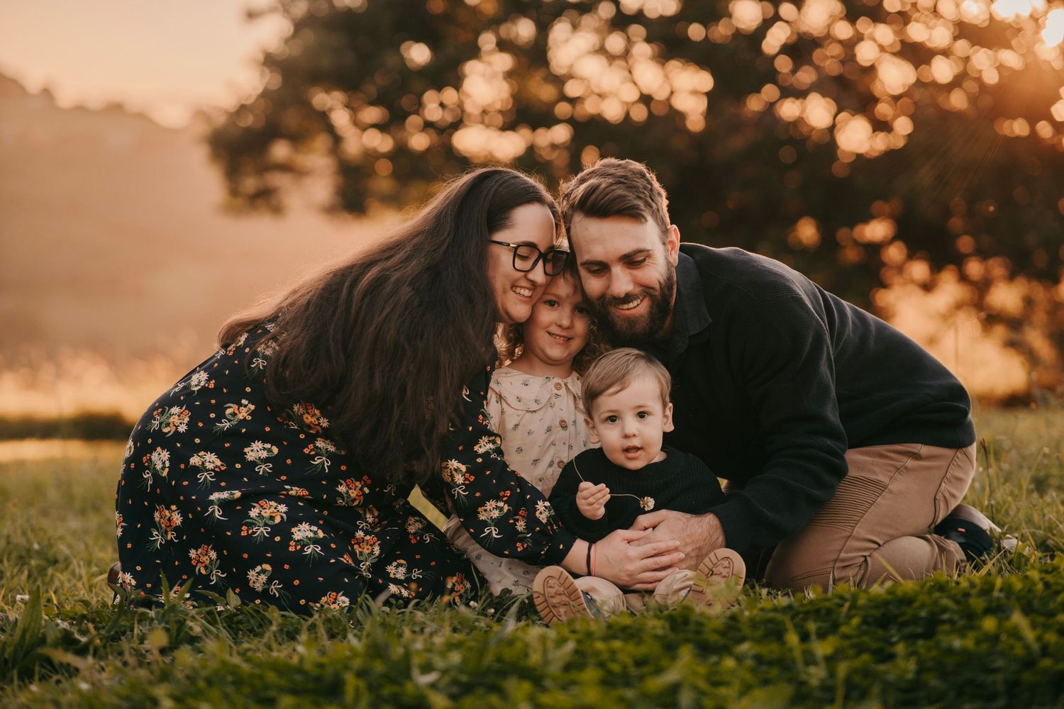 Happy family of four sitting on green grass during golden sunset, parents cuddling two young children outdoors.
