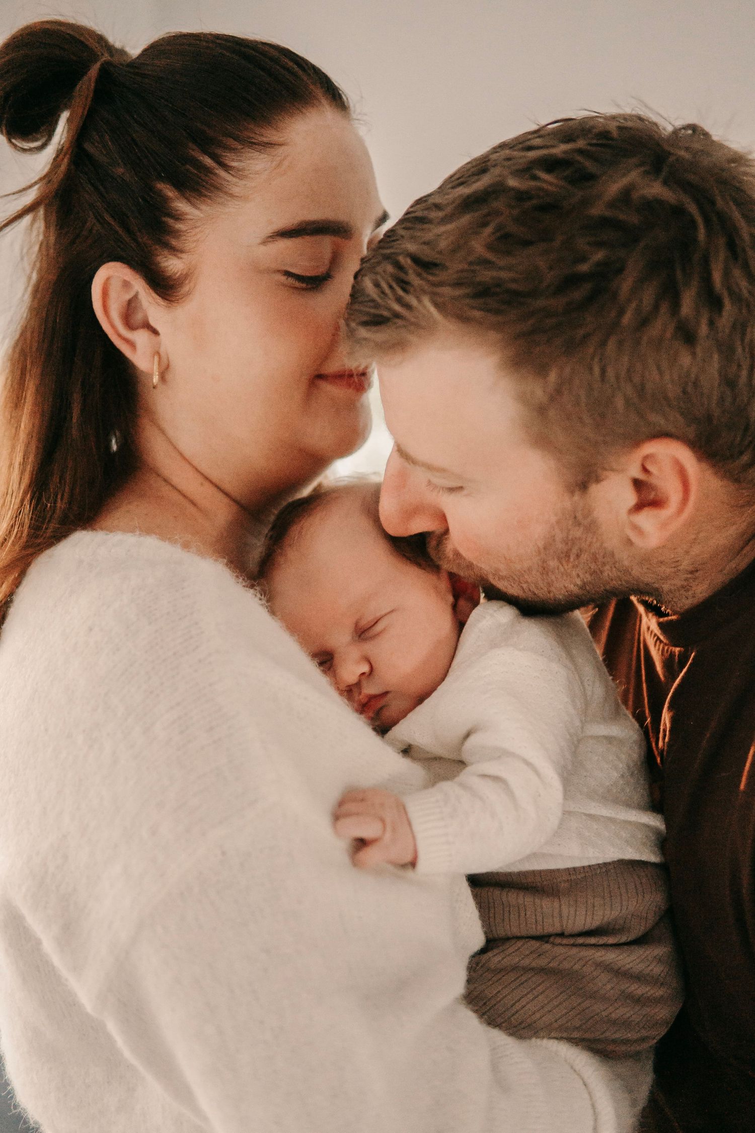 Loving parents touching foreheads with sleeping newborn baby in warm intimate portrait.