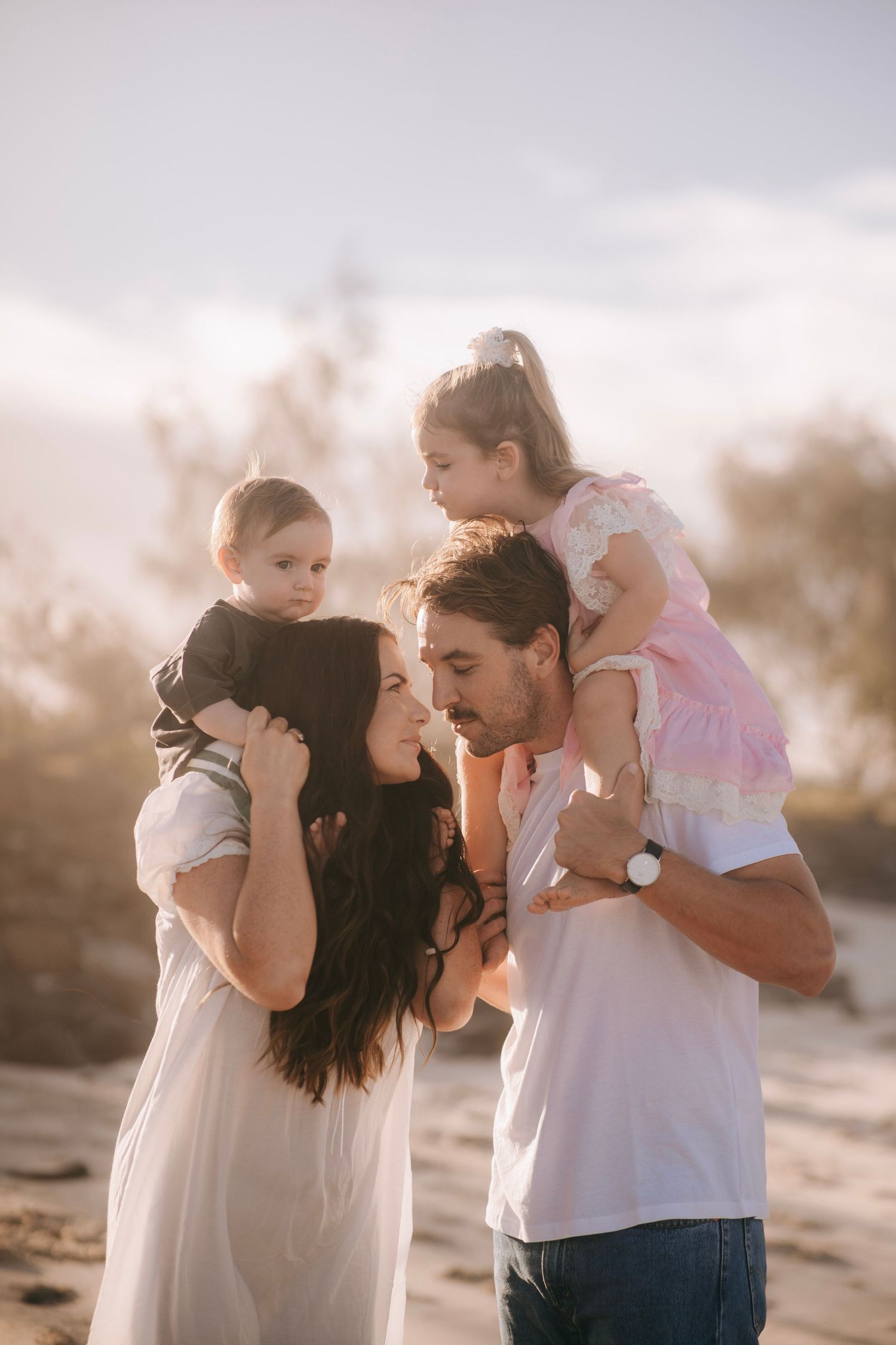 Loving family portrait at sunset with parents holding two young children on sandy dunes in warm light.