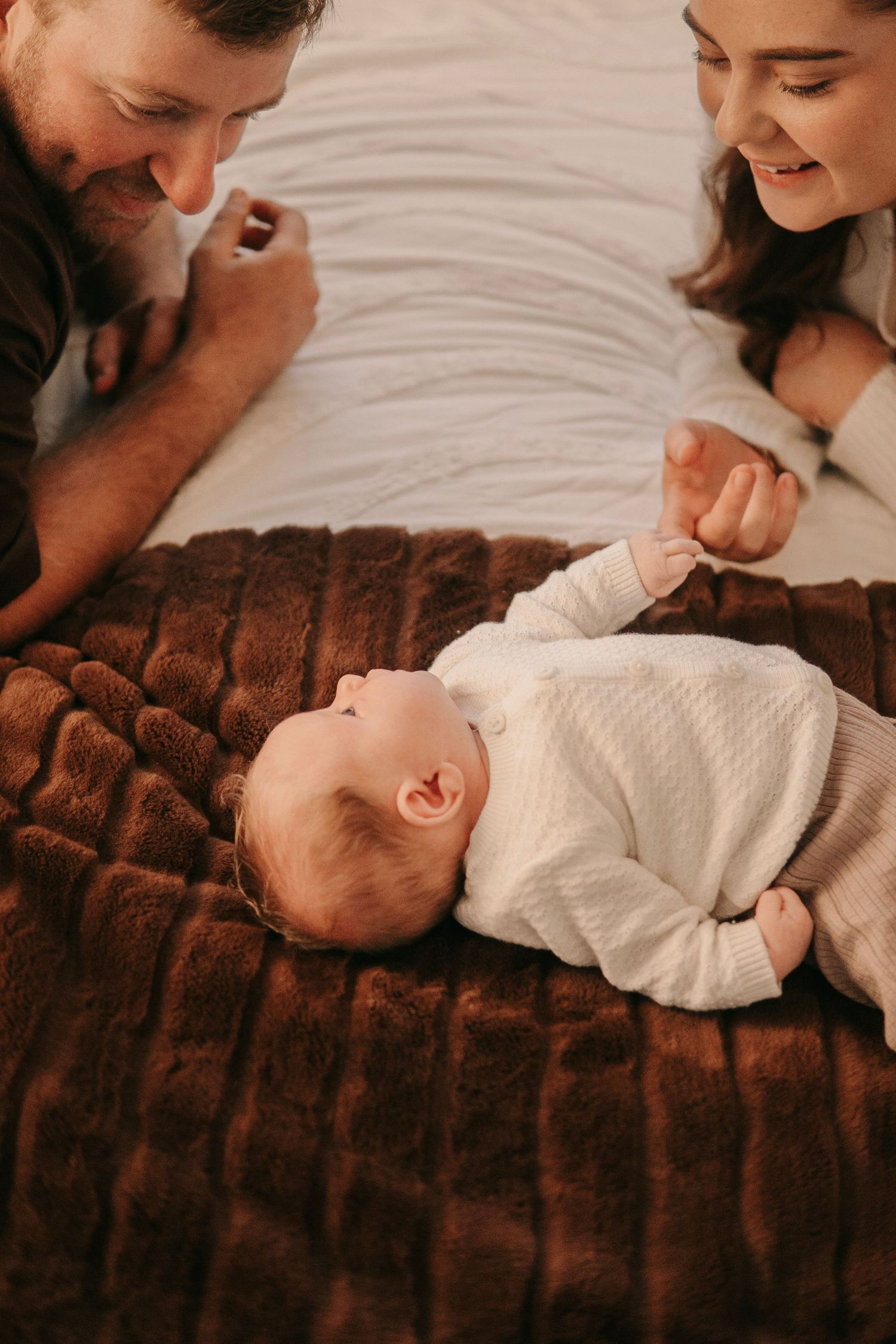 Happy parents smiling at their newborn baby lying on a brown fur blanket.