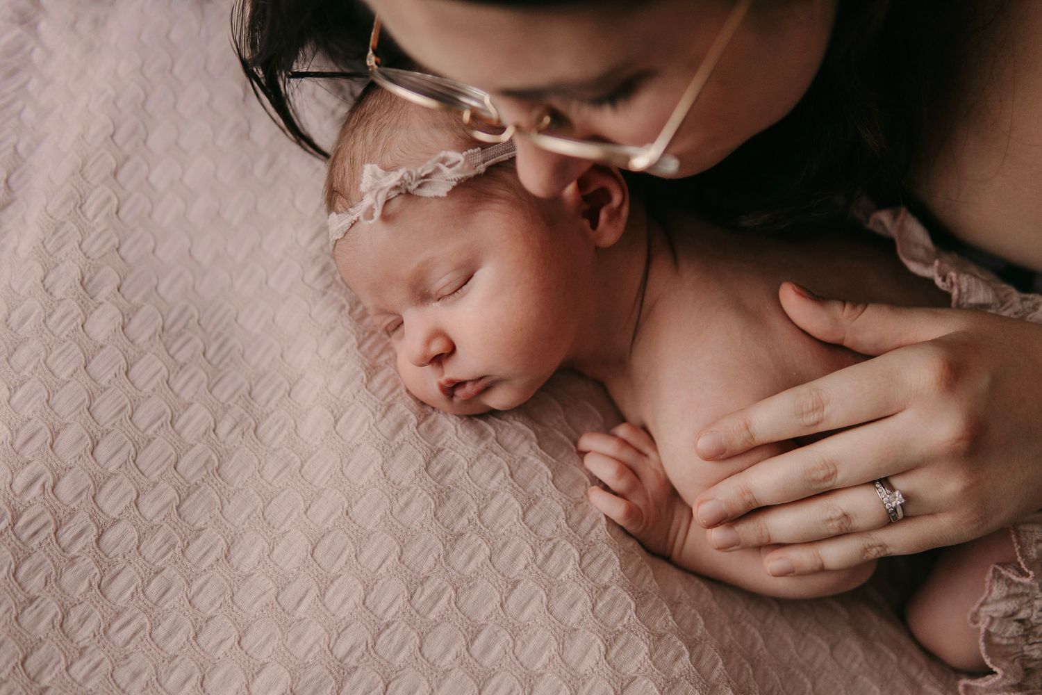A mother tenderly kisses her sleeping newborn baby wearing a delicate floral headband on a soft pink blanket.
