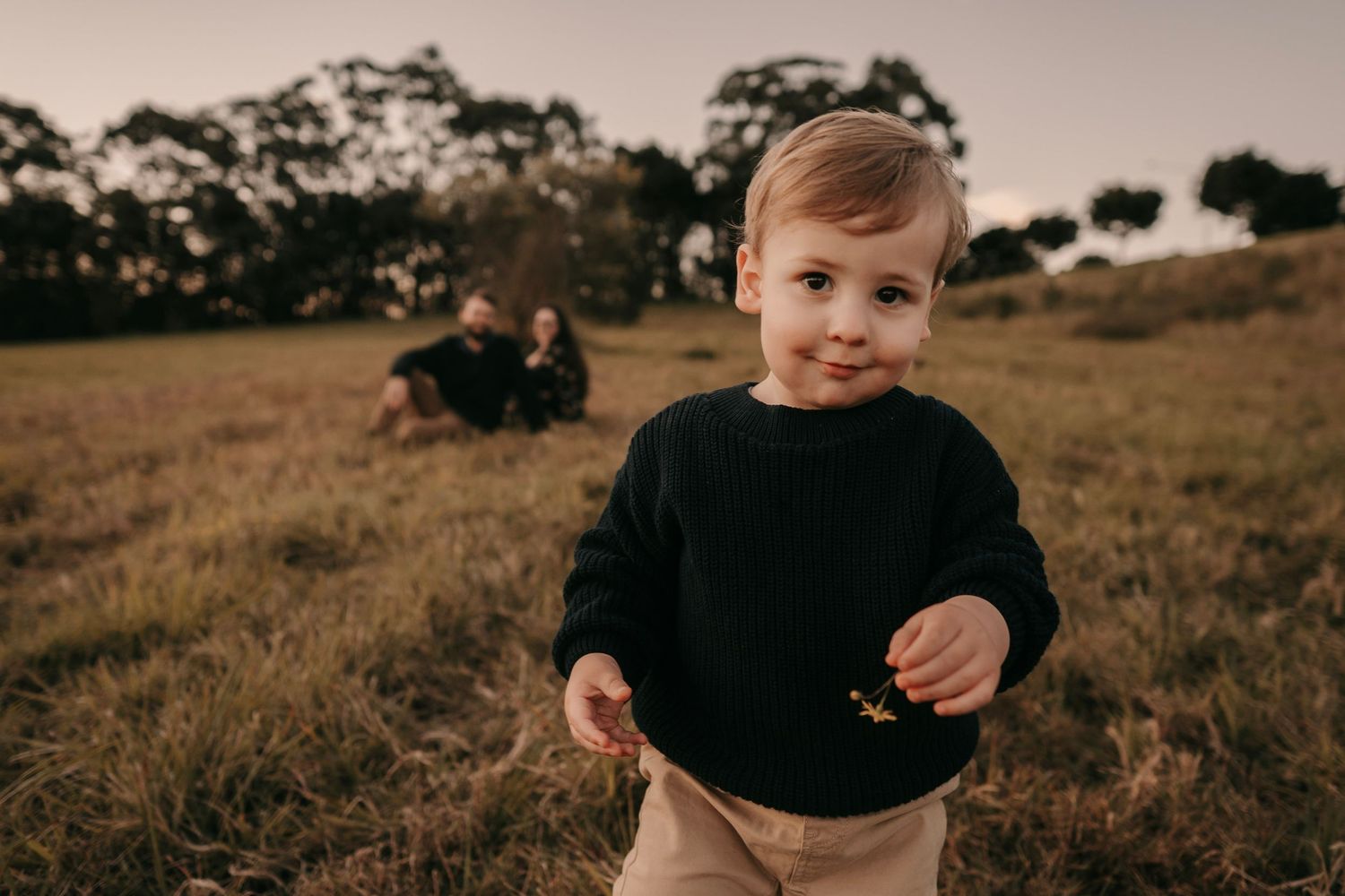 An adorable toddler boy in a black sweater stands in an open field at golden hour.