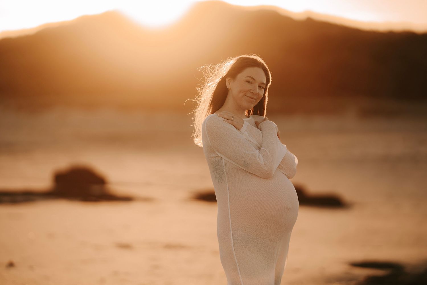 Pregnant woman in white dress glowing in golden sunset light on a sandy beach.