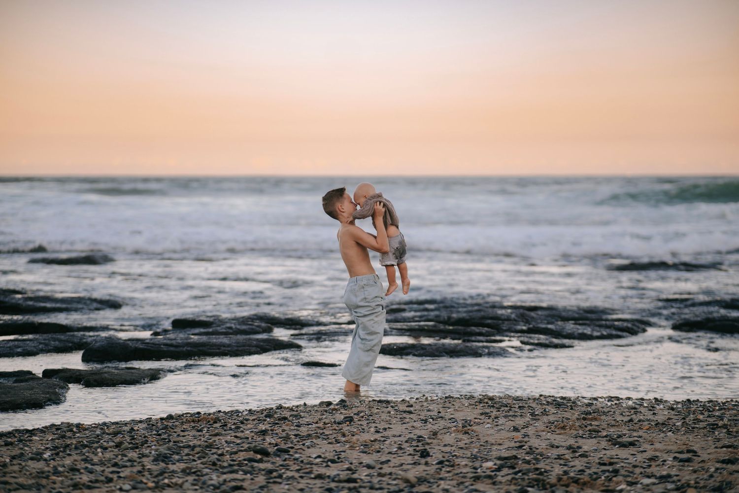 Father lifting baby joyfully on rocky beach at sunset with ocean waves behind.