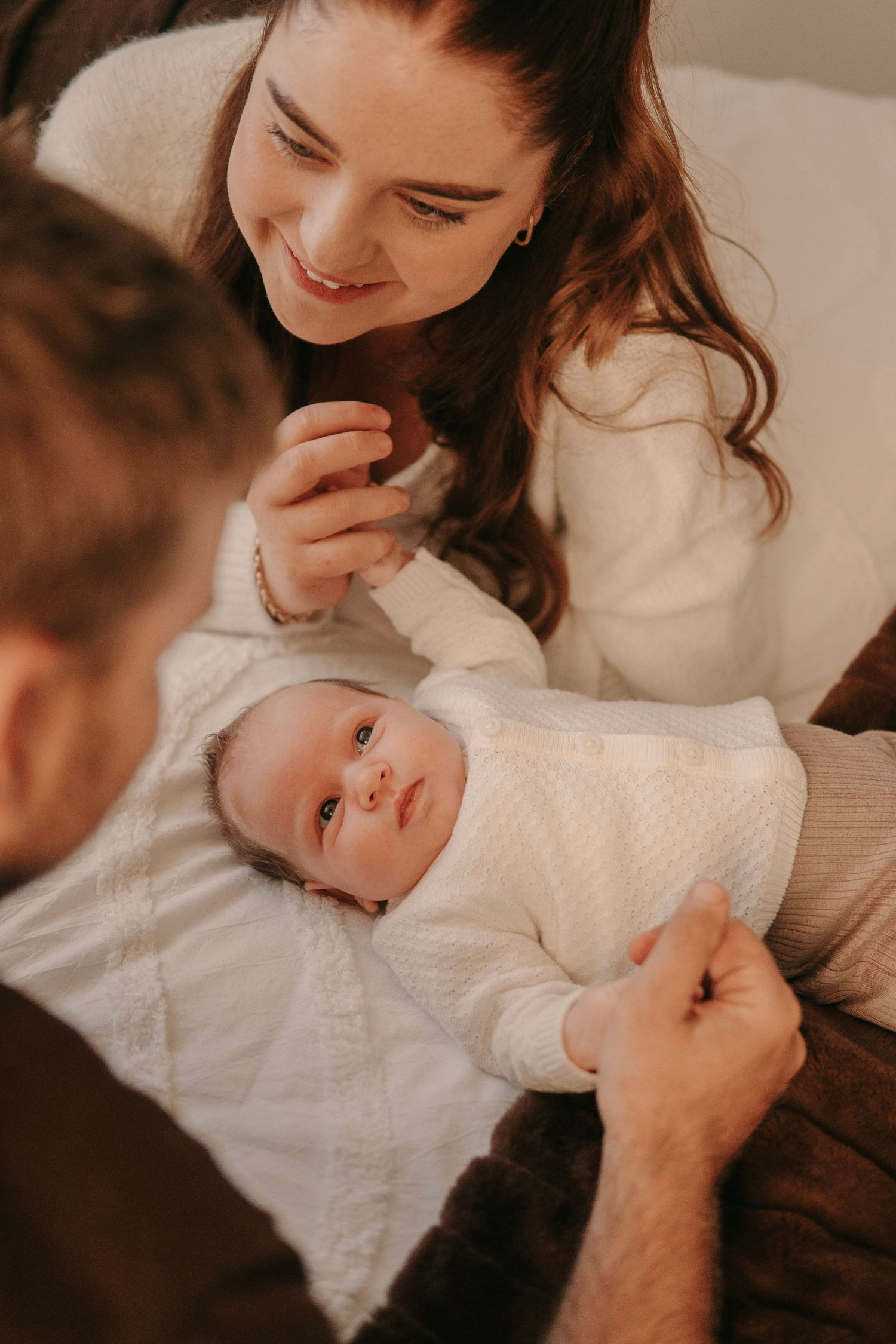 Smiling mother with long brown hair looking at her newborn baby in a white knit outfit.