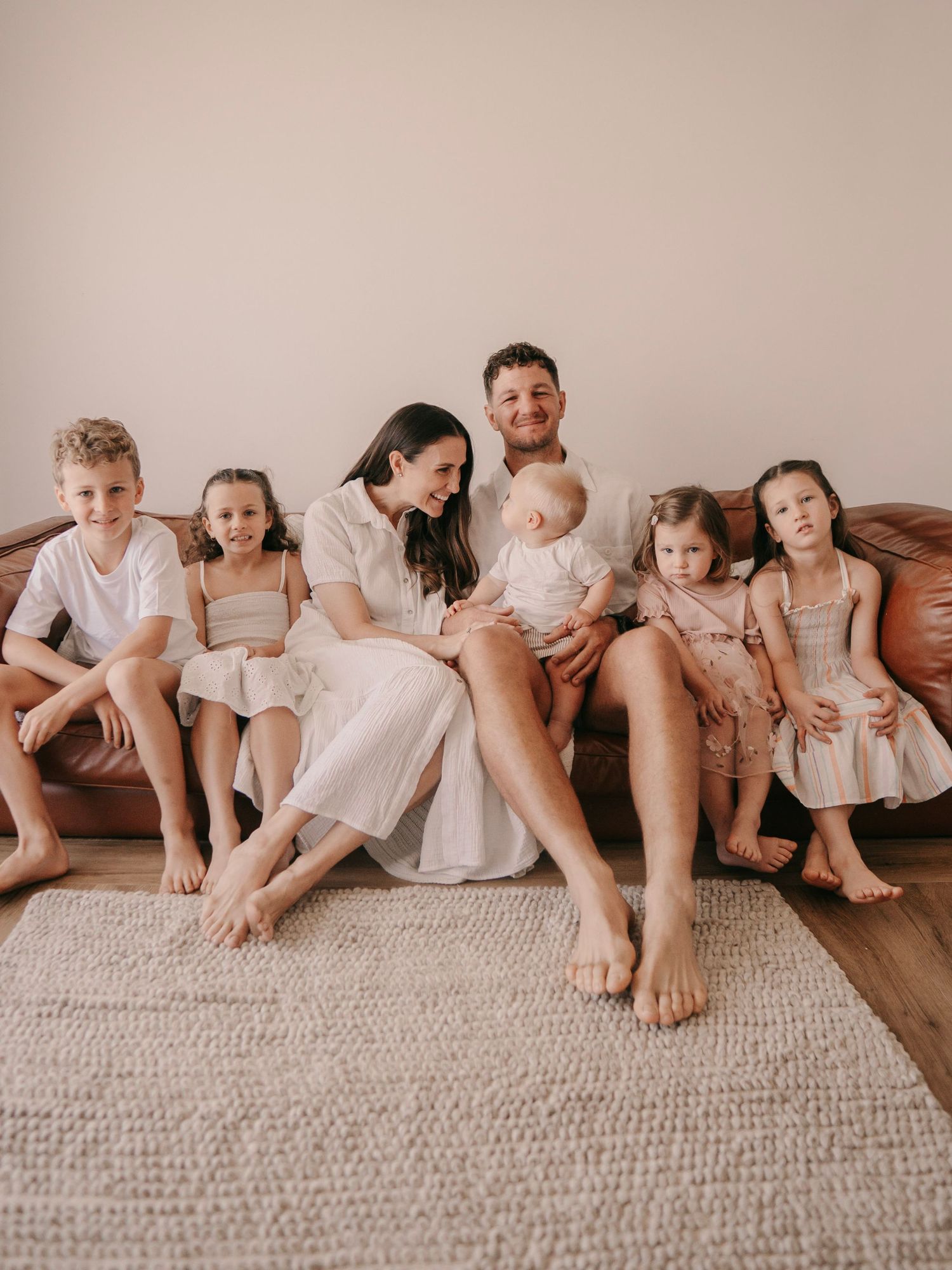 A happy family of seven sits together on a brown leather couch, dressed in neutral tones for a cozy portrait.
