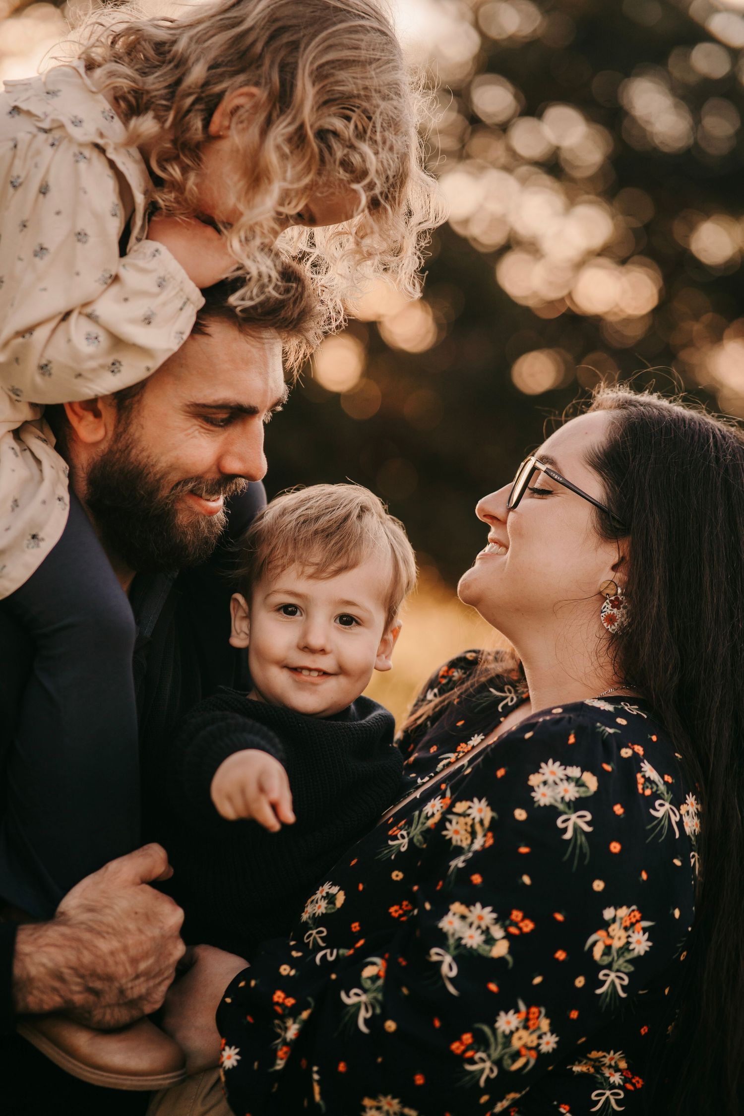 A bearded father and mother with glasses share a joyful moment with their two young children outdoors at golden hour.