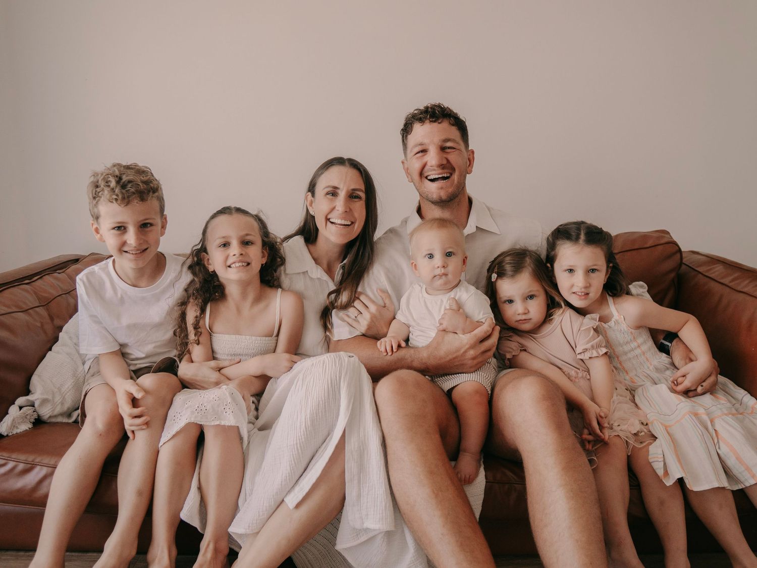Happy family of seven posing on a brown leather couch, parents with five young children in neutral tones.