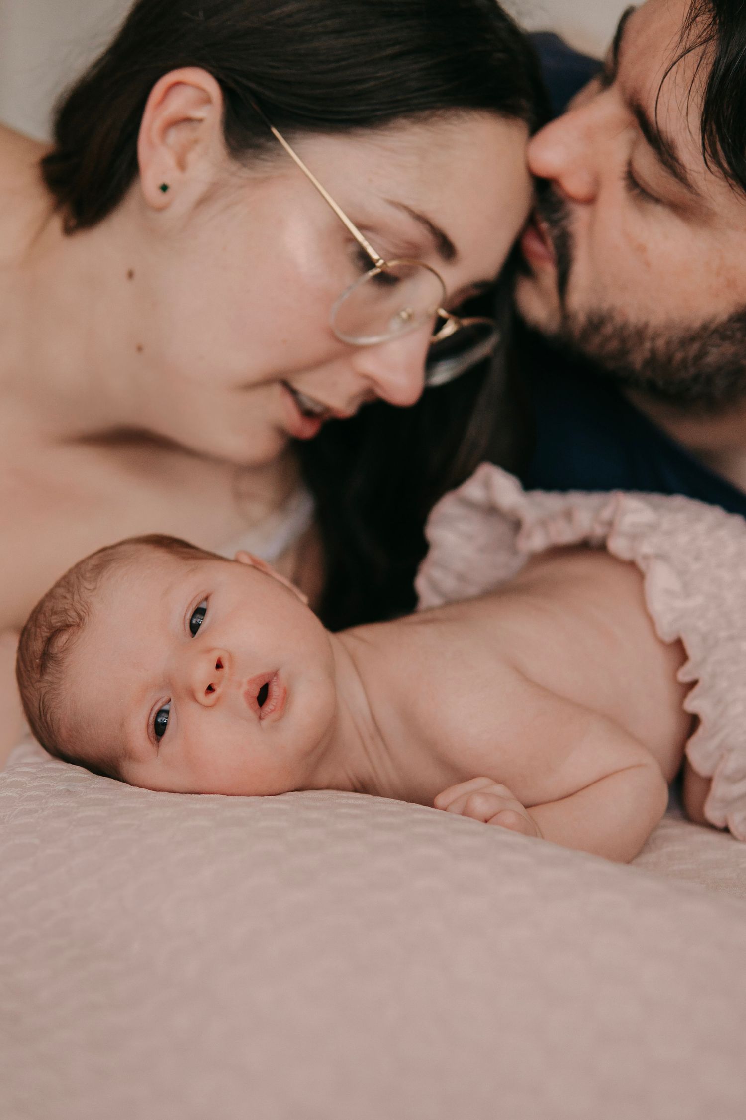 New parents lovingly gazing at their alert newborn baby lying on a soft white bed.